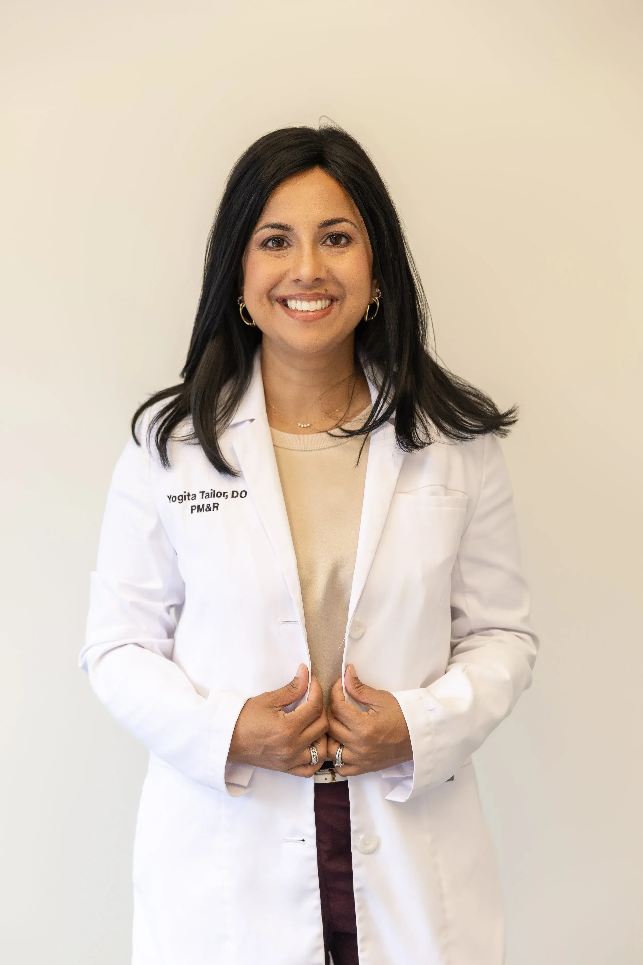 Dr. Yogita Tailor wearing a white medical coat, smiling, holding her coat closed with both hands, standing against a plain beige wall.