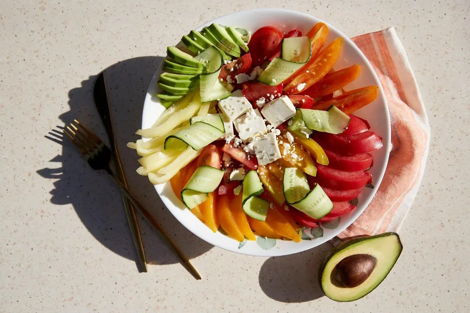 A white bowl of sliced colorful vegetables, including avocado, cherry tomatoes, bell peppers, cucumber, and cheese, on a speckled white surface with a halved avocado and a folded cloth napkin nearby.