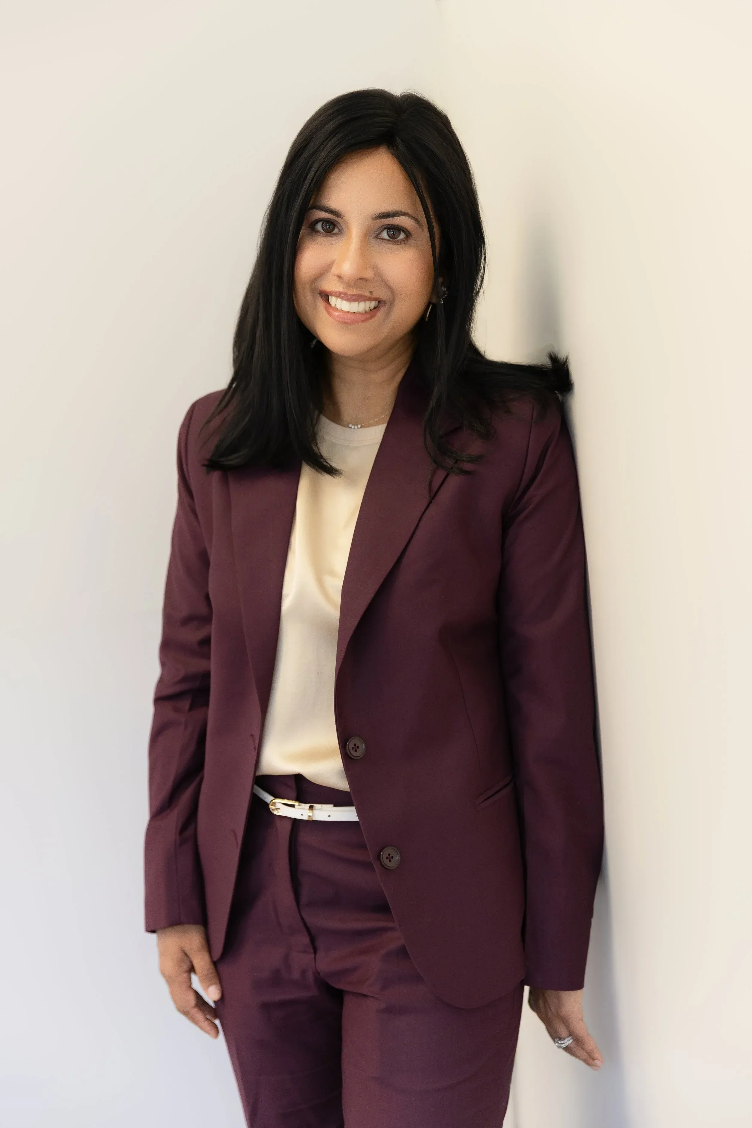 Dr. Yogita Tailor with black hair wearing a burgundy suit and light-colored blouse standing against a plain white wall, smiling at the camera.