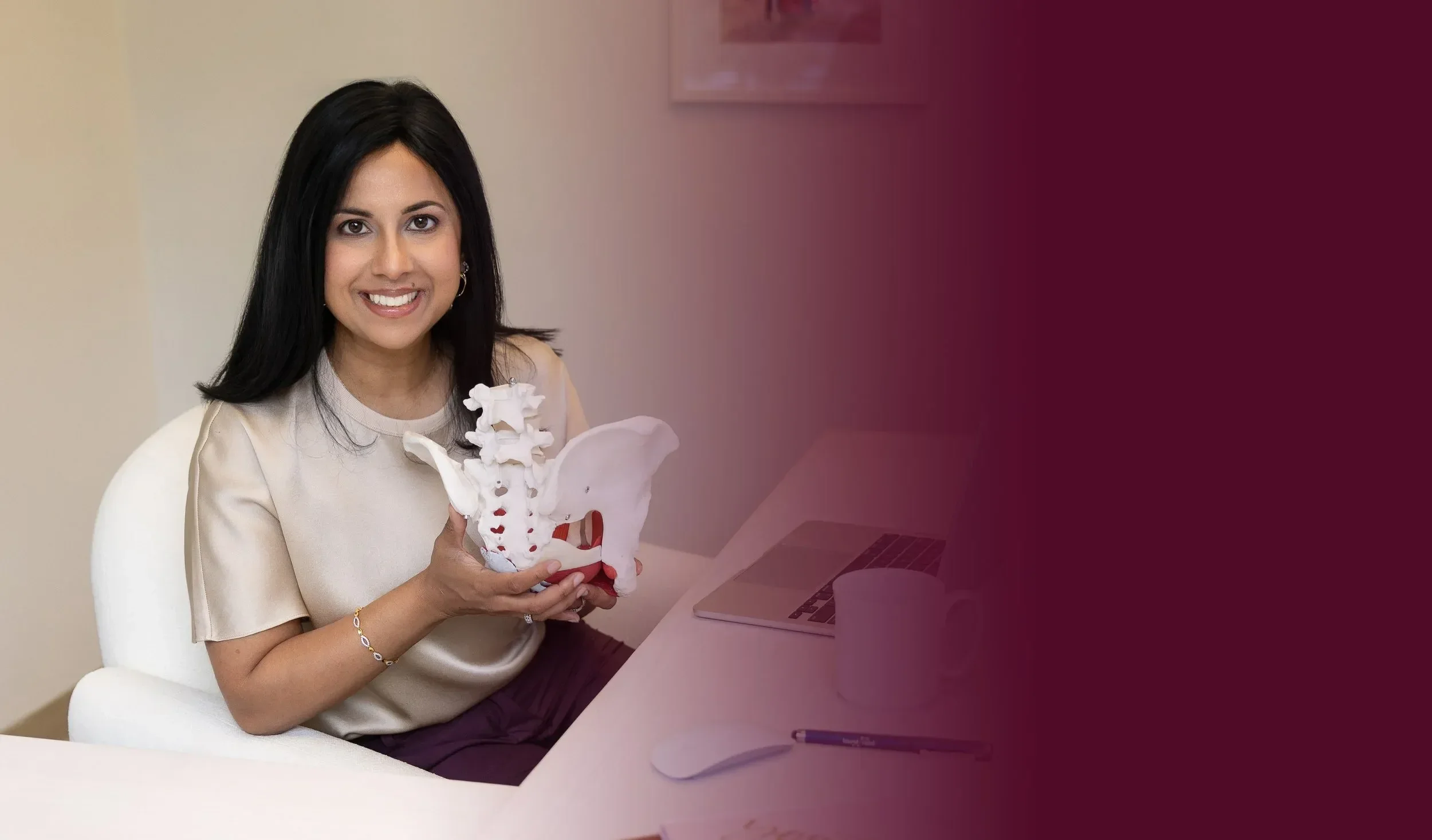 Dr. Yogita Tailor with black hair smiling and holding a model of a human pelvis, sitting at a desk with a laptop and a mouse.