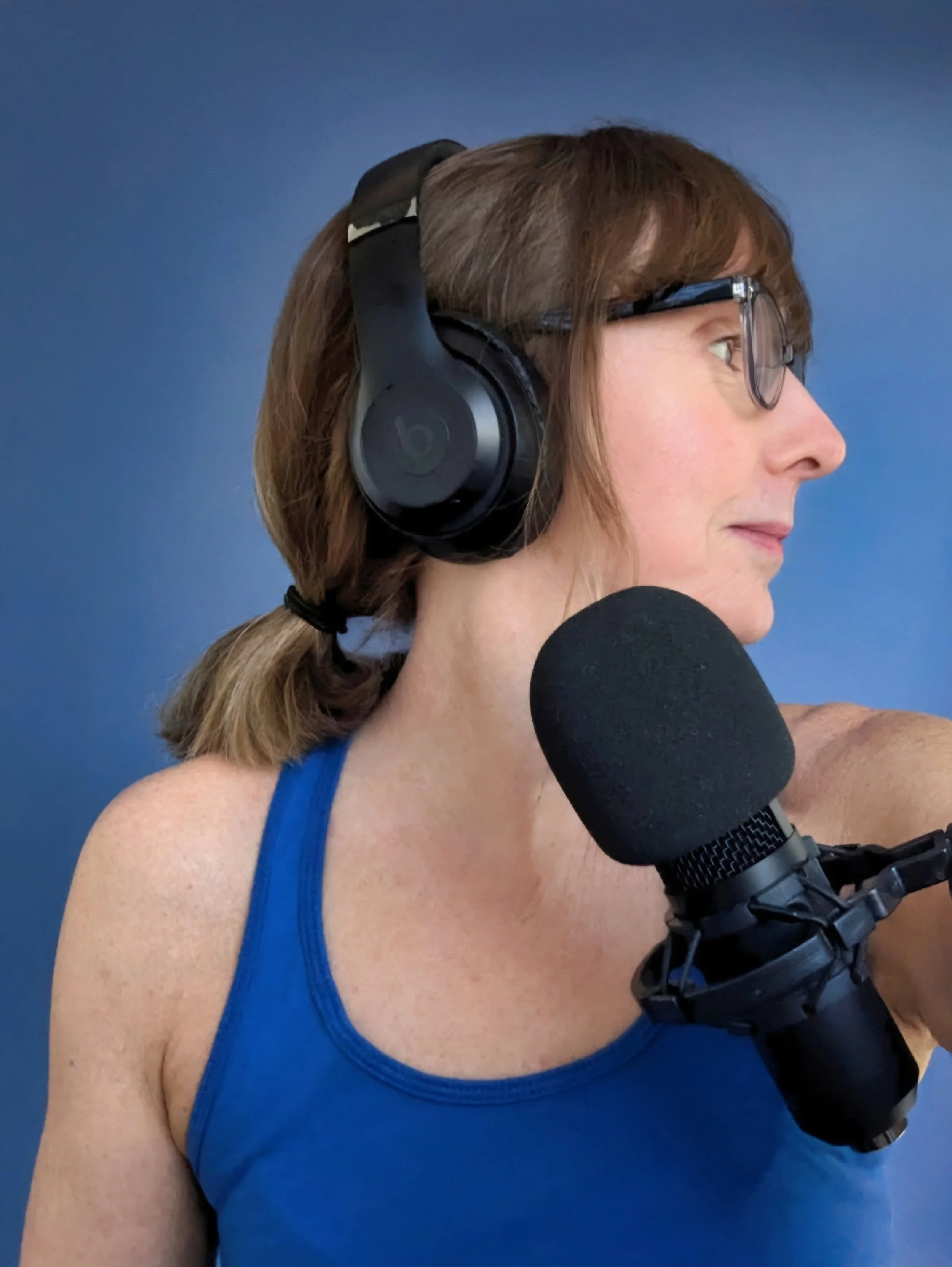 A woman with brown hair tied in a ponytail, wearing glasses, a blue tank top, black headphones, and a microphone setup, looking to the right against a plain blue background.