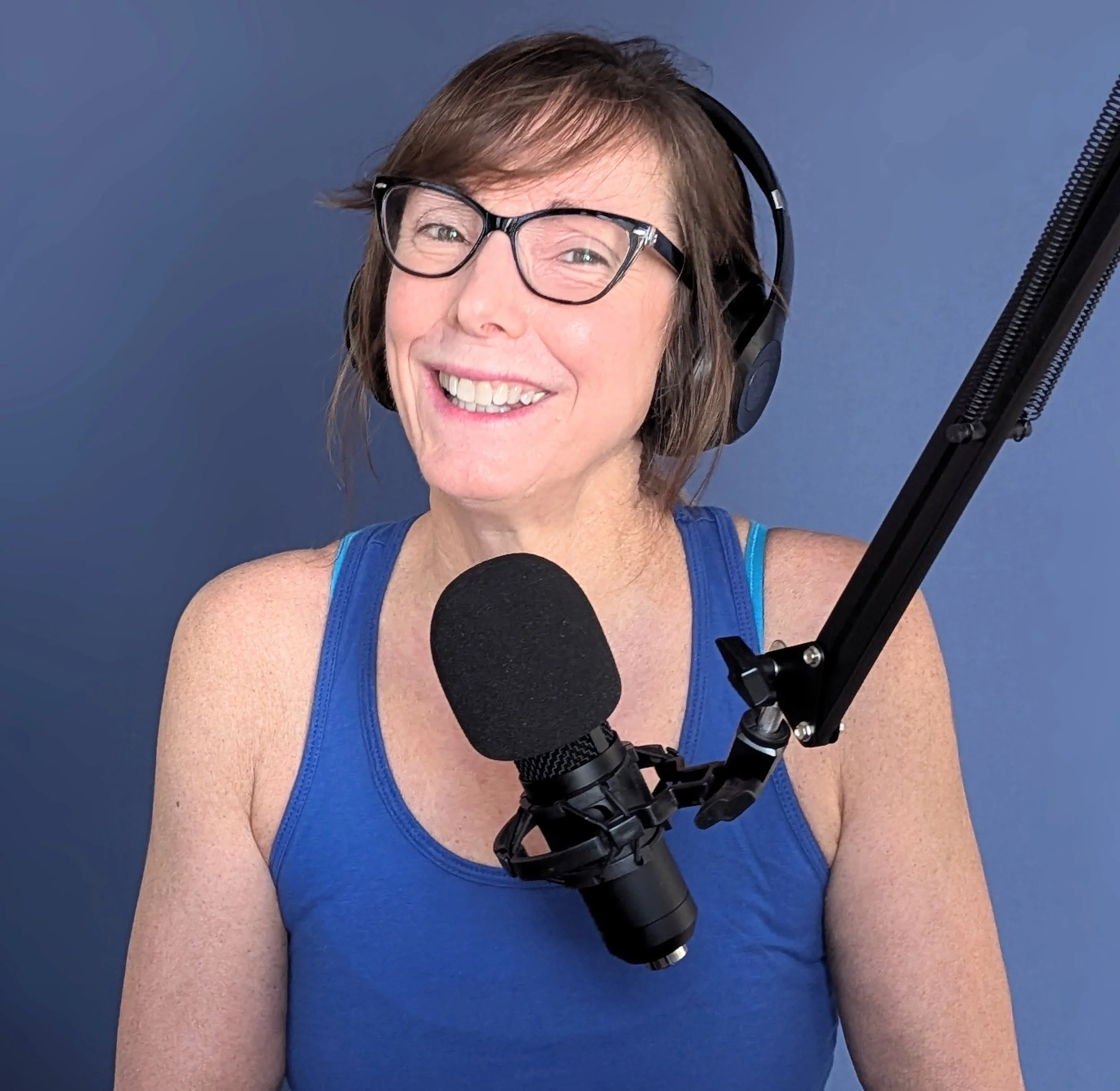 Smiling woman wearing glasses and headphones speaking into a microphone against a blue background.