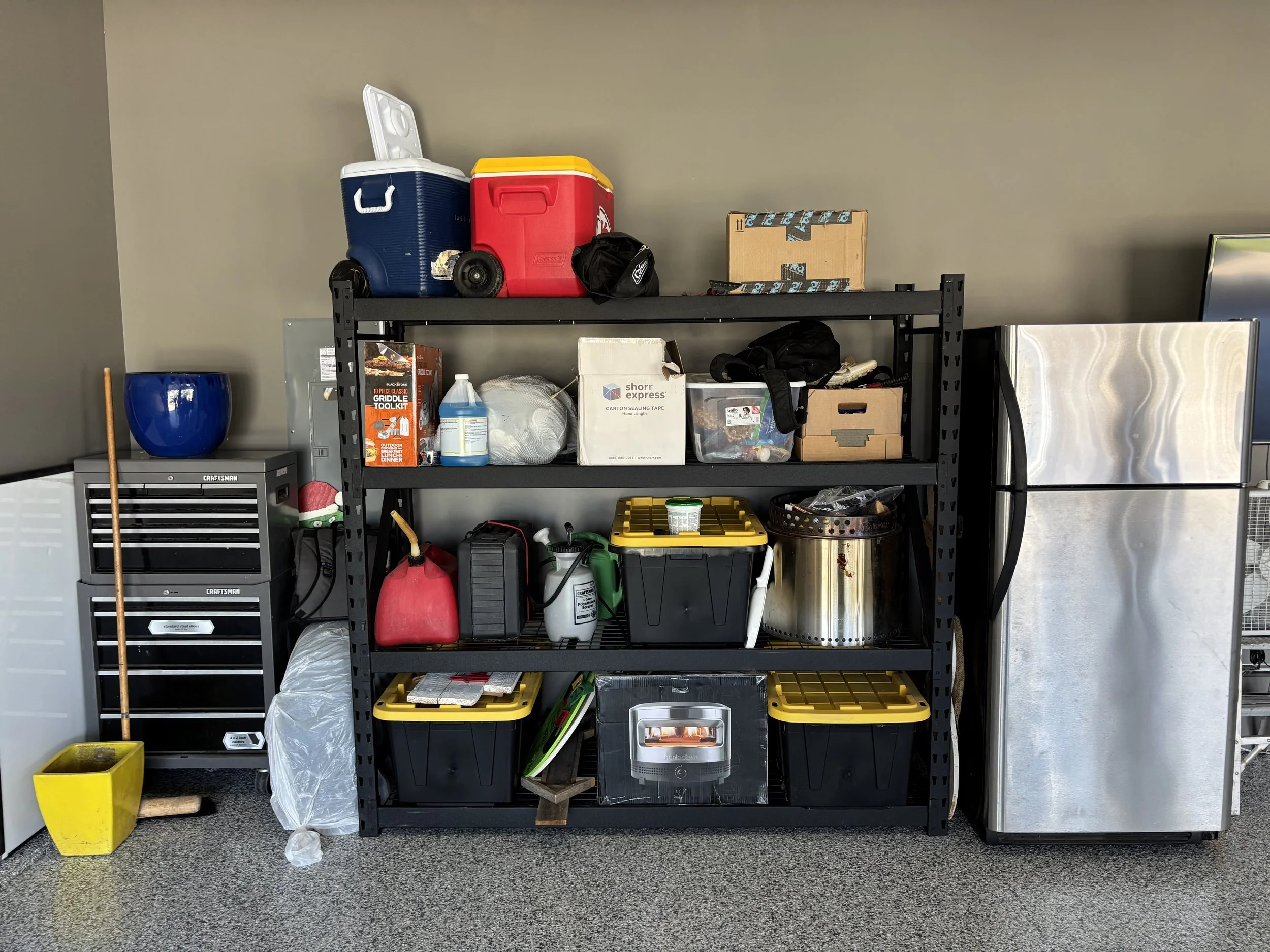 Garage storage shelf filled with various tools, containers, and supplies, next to a refrigerator, with a mop and yellow plant pot on the floor.