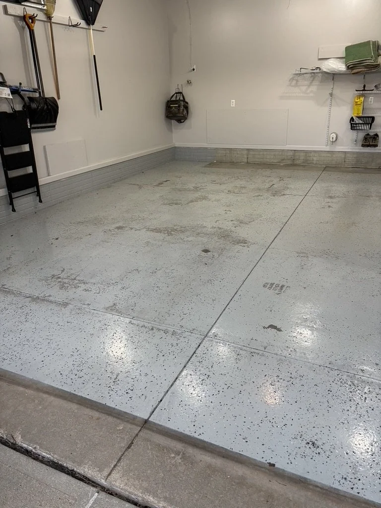 Empty garage with a concrete floor, some tools hanging on the wall, and storage shelves with a few items.