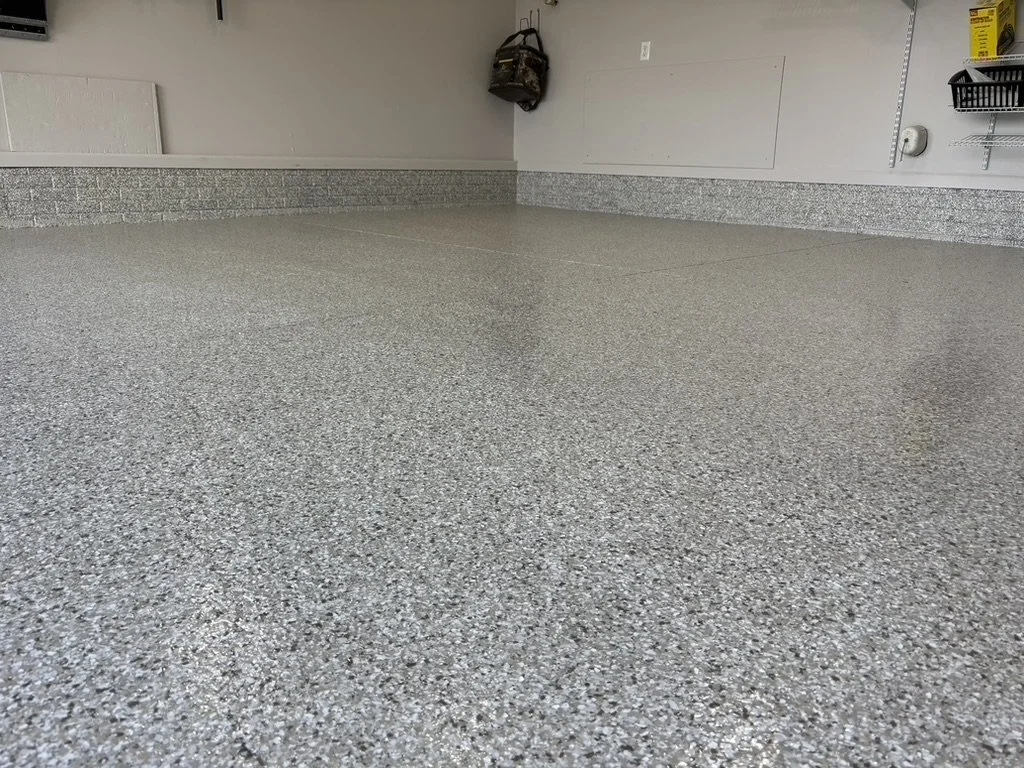 Empty garage with speckled gray epoxy flooring, white walls, and a black storage basket on a wire shelf.