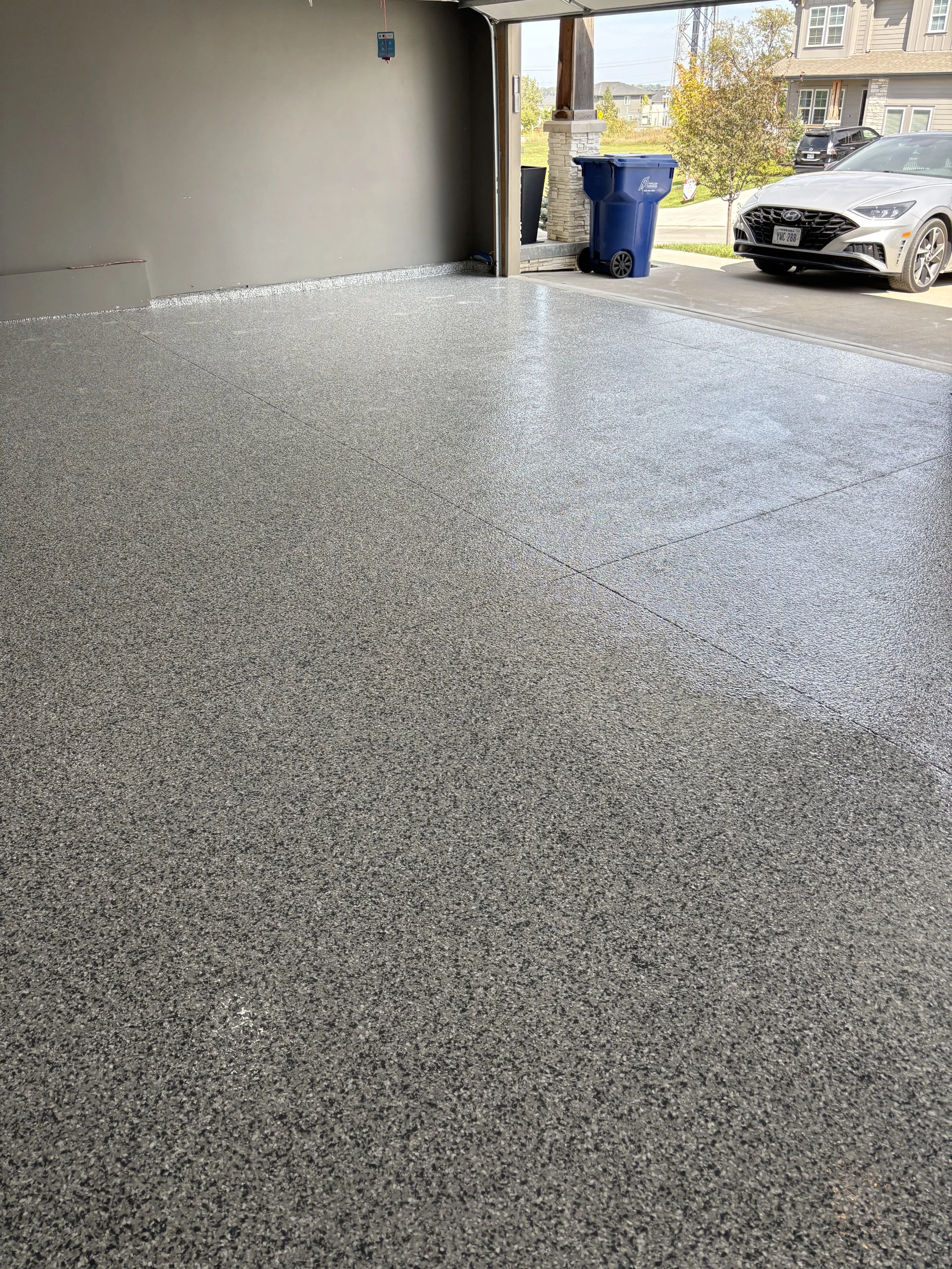 Empty garage with textured epoxy floor, a view of driveway outside where a white car is parked, a blue trash bin, and residential houses in the background.