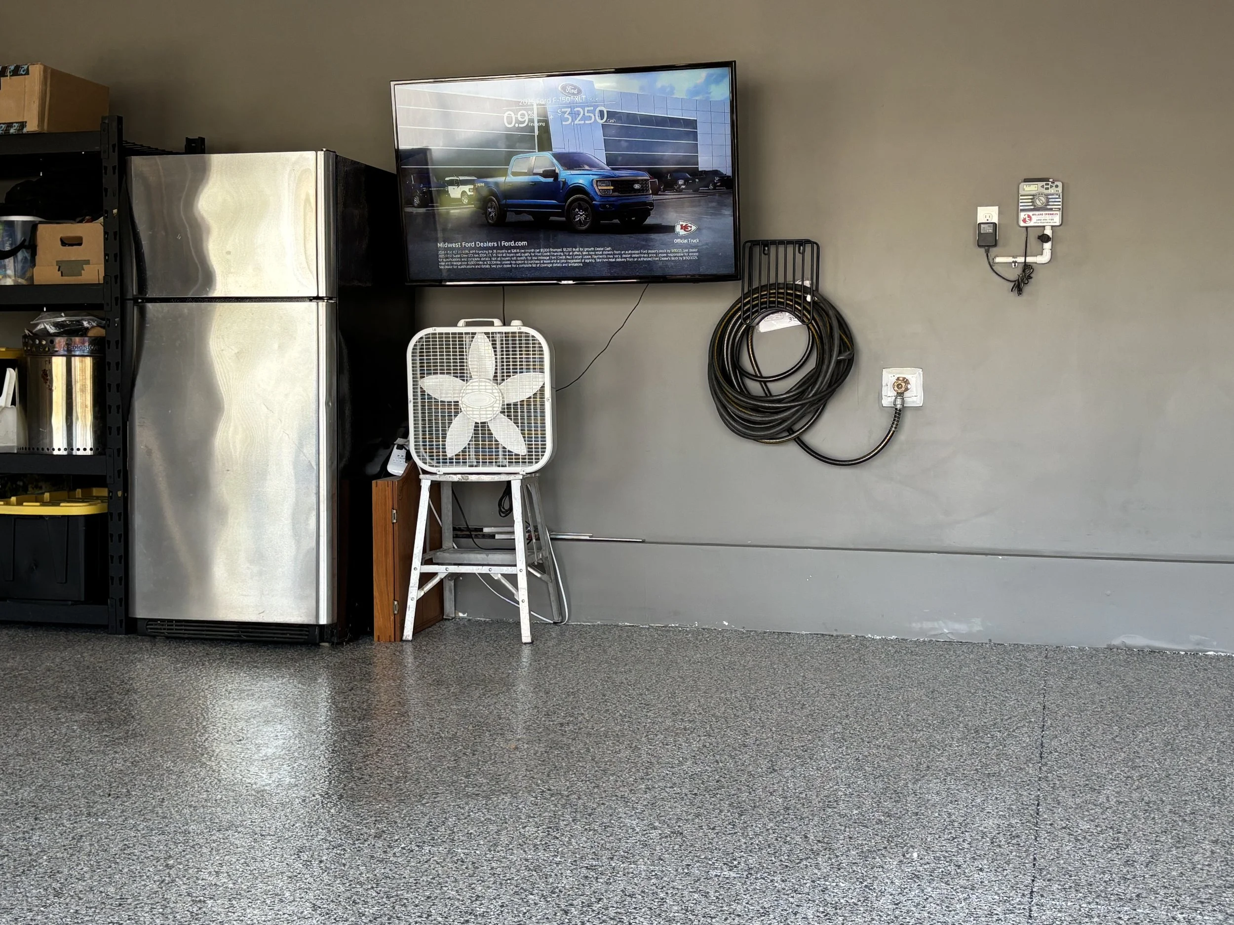 A garage or workshop wall with a mounted television screen showing an advertisement for a vehicle, a folding fan on a small stand, a black coiled cable mounted on the wall, and electrical outlets.