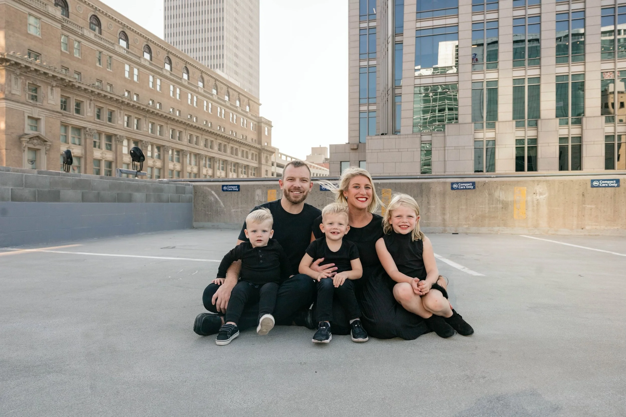 A family of five sitting on the ground of an outdoor parking garage, with city buildings and skyscrapers in the background, all smiling and dressed in black.