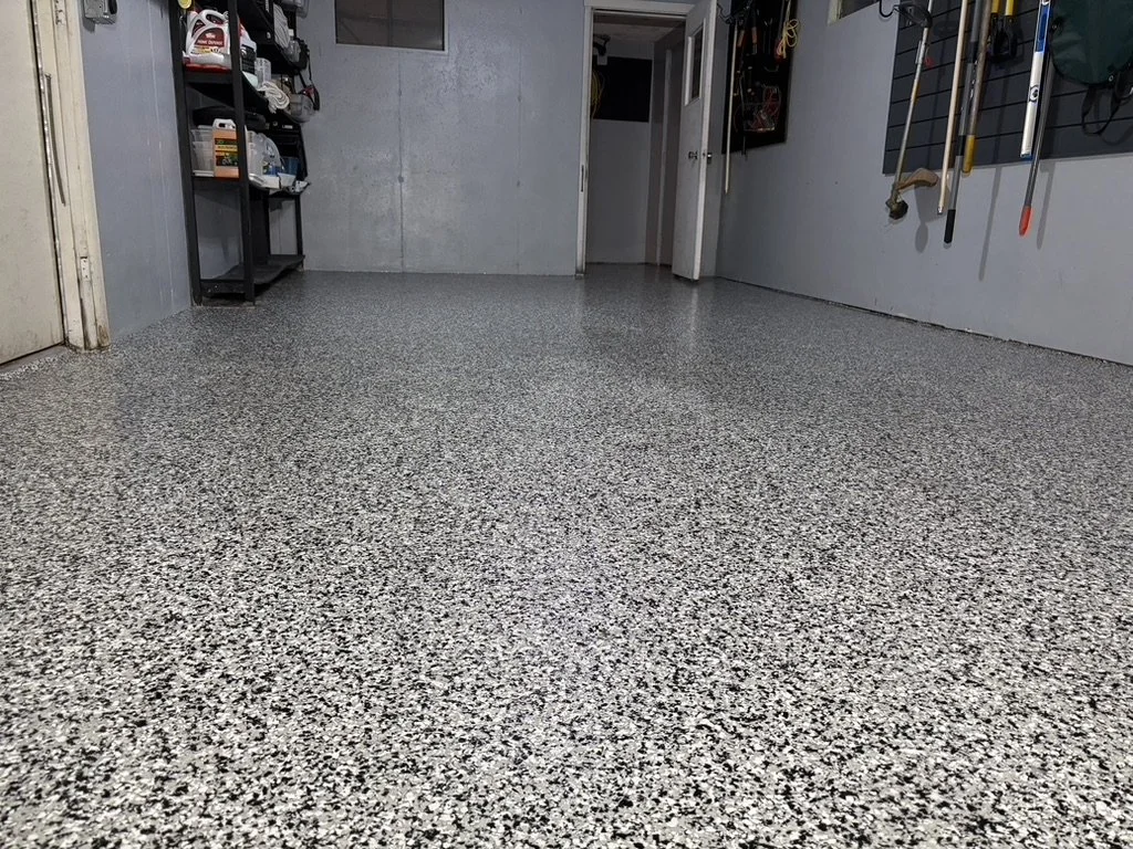 Empty garage with a speckled epoxy floor, gray walls, and shelves with supplies on the left side, tools and equipment hanging on the right wall, and a door at the back.