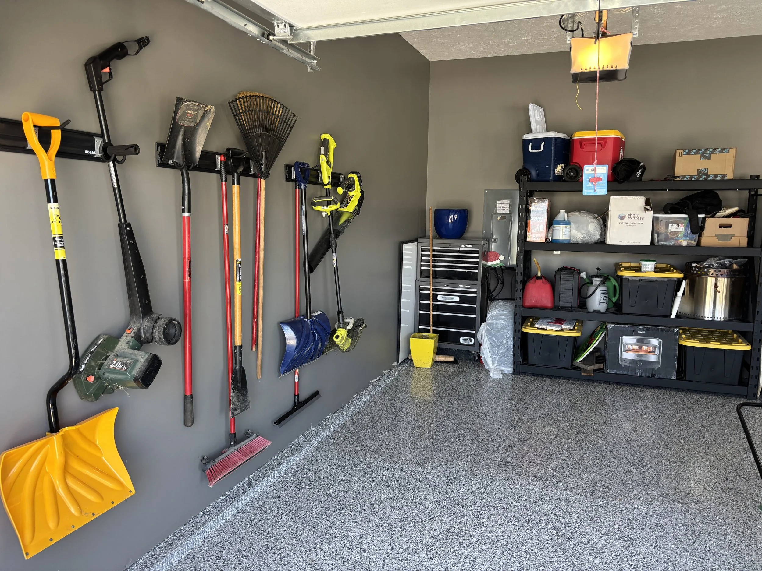 Garage with tools hanging on a gray wall, including a snow shovel, leaf blower, rake, and snow blower, with shelves holding coolers, containers, and miscellaneous items on the right side.