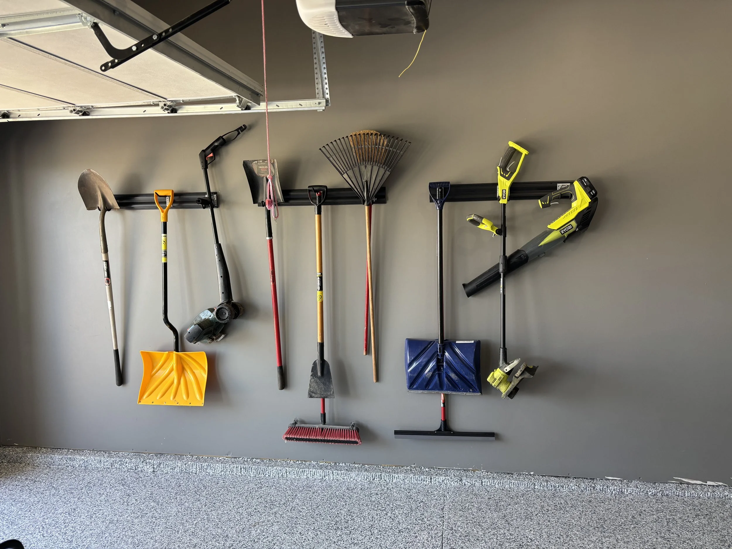 Garage wall with various gardening tools hanging, including shovels, a rake, a snow shovel, and a leaf blower.
