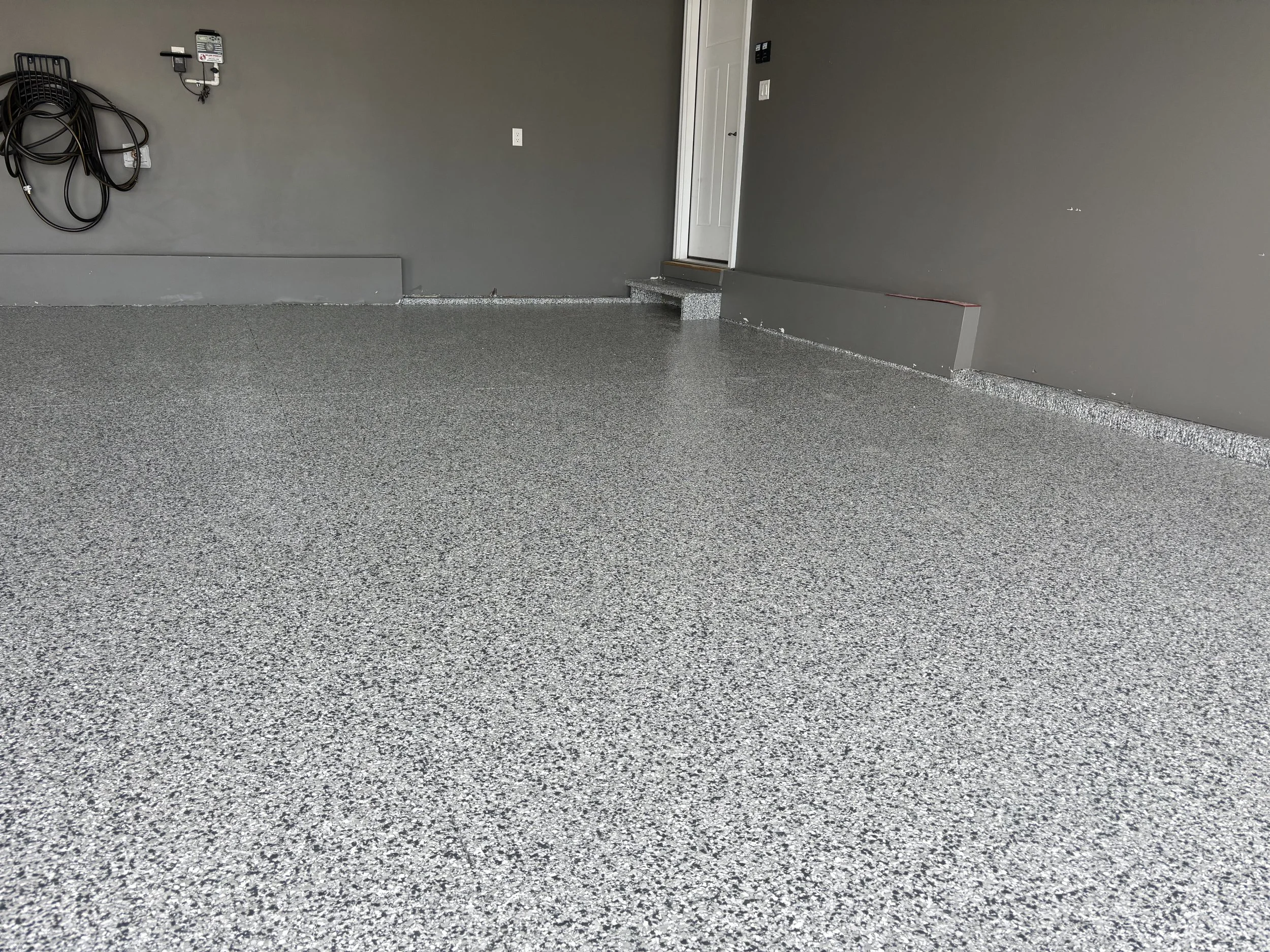 Empty garage with a gray speckled epoxy floor, gray walls, and a closed white door. Hoses and electrical outlets are mounted on the wall.