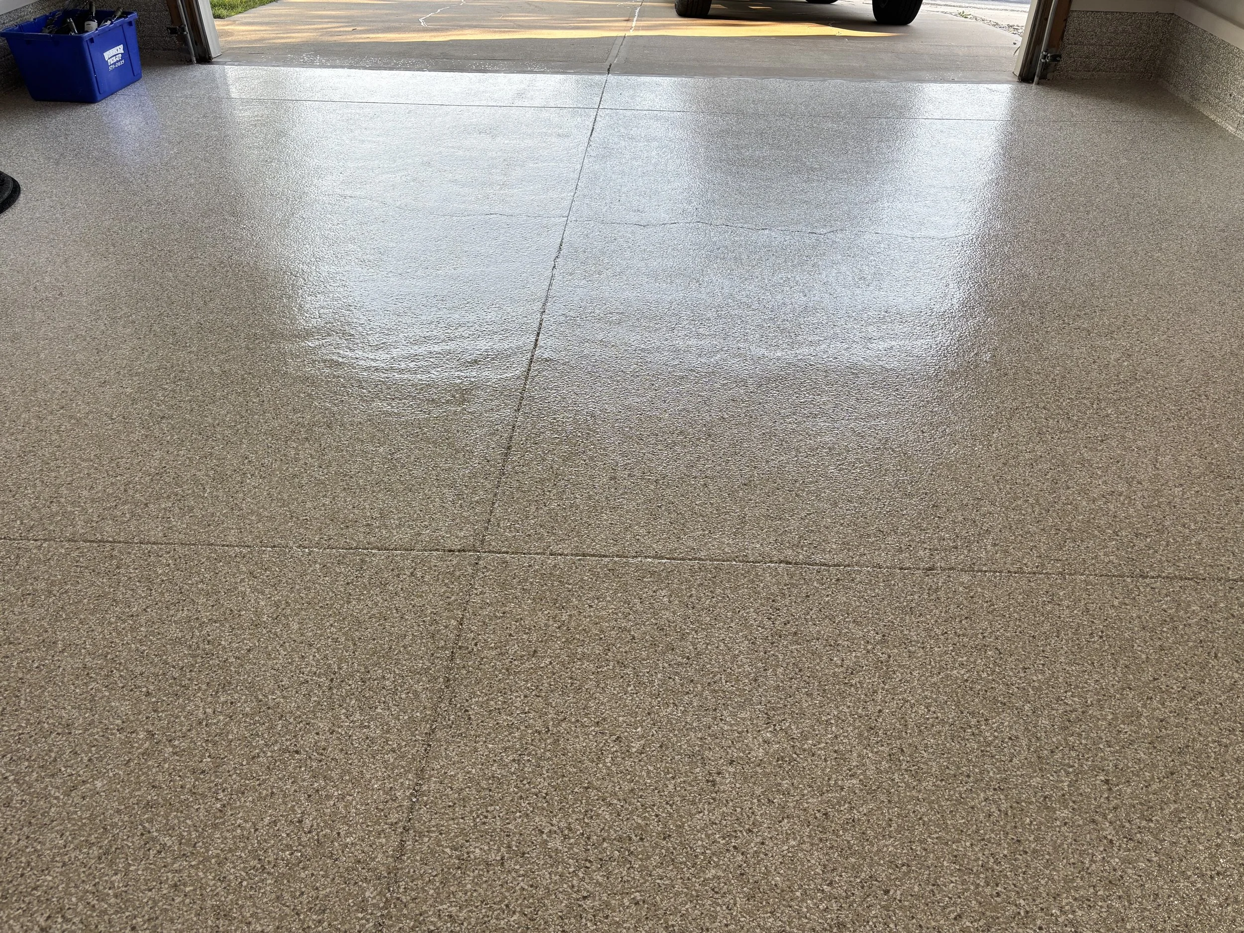 Clean, shiny garage floor with a section open to the outside driveway, a blue recycling bin and a car tire visible.