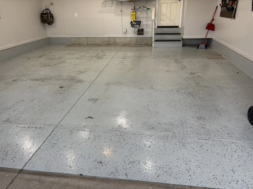 Empty garage with a concrete floor, some stains, and a few cracks. There are shelves and tools on the far wall and a red broom leaning against the wall.