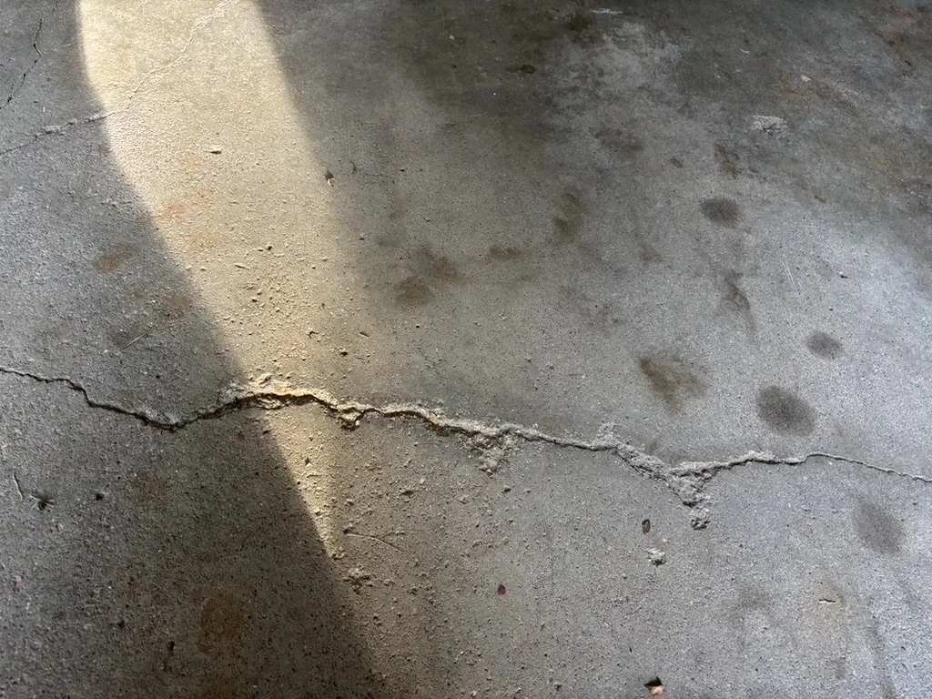 Concrete floor with a prominent crack running across it and some dark stains, with a shadow cast on part of the floor.
