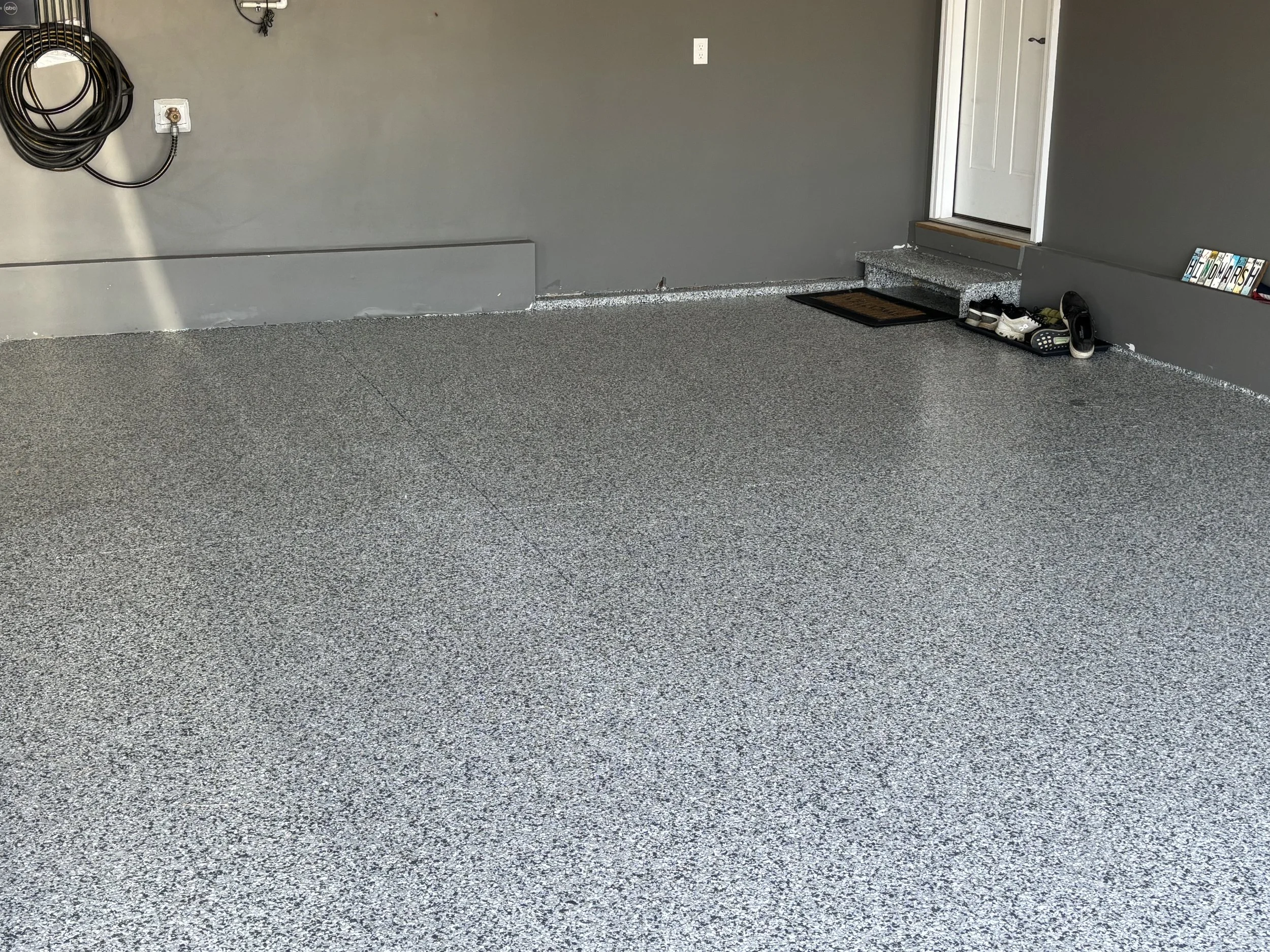 Empty garage with gray walls, speckled gray flooring, a coat of paint near the door, and a few pairs of shoes near the entrance.