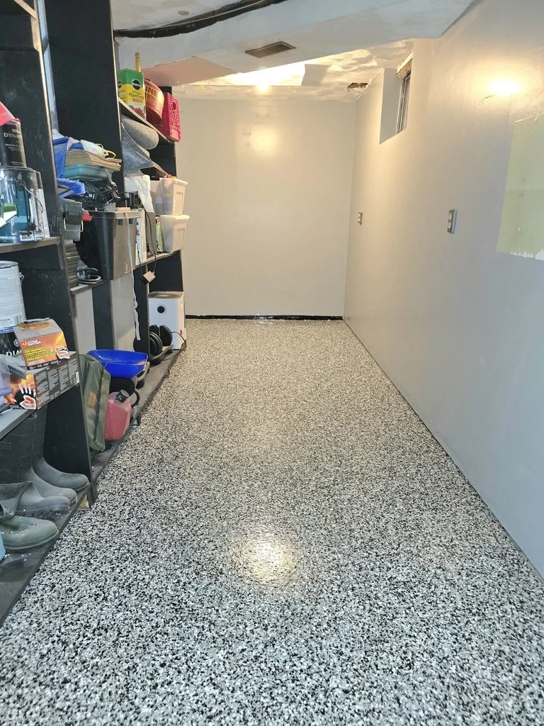 Empty laundry or utility room with storage shelves on the left and a plain wall with a small window on the right. The floor has a speckled gray and white pattern.