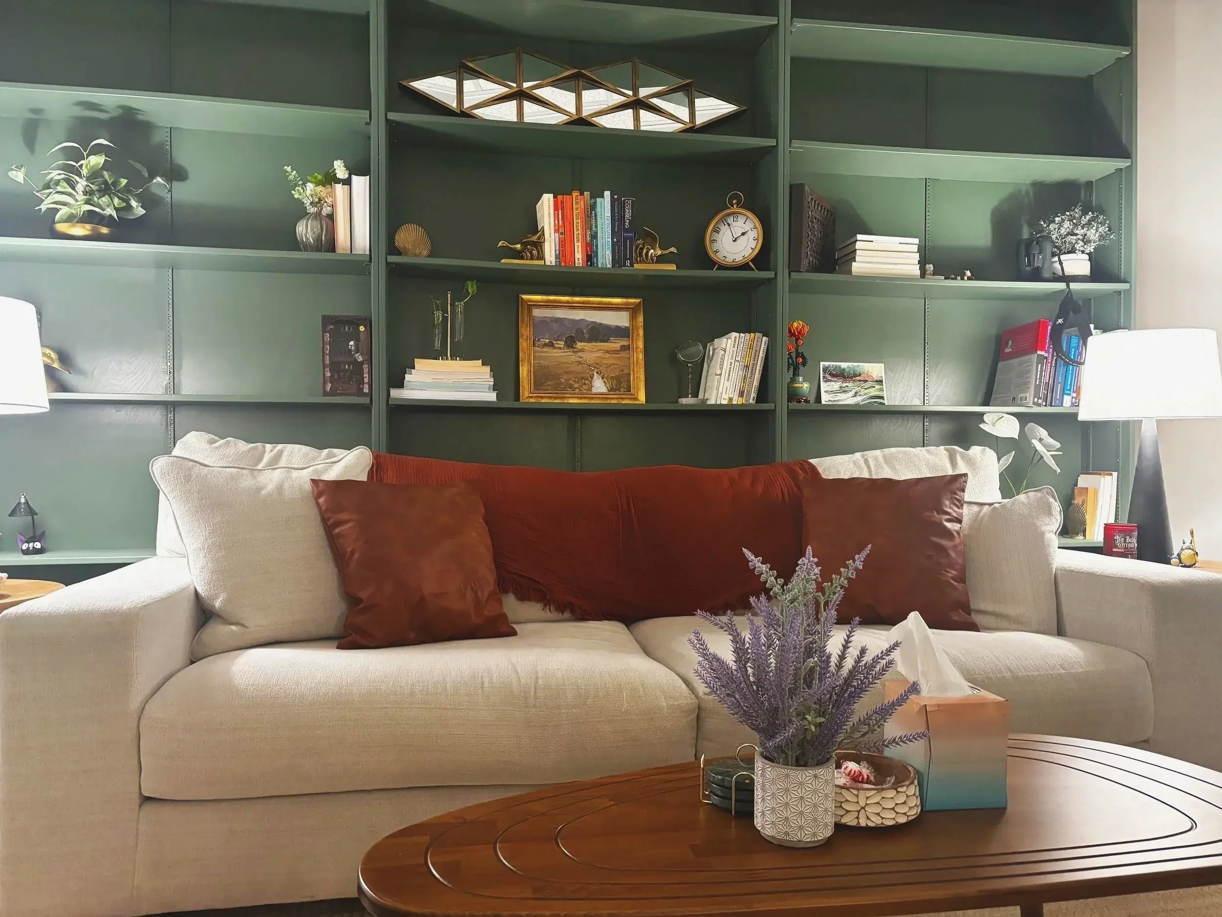 A beige sofa with brown pillows in front of a green bookshelf filled with books, plants, and decorative items, with a lamp on each side.