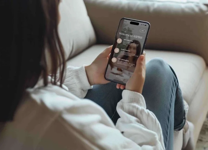 Woman sitting on a couch using her phone, viewing the Aspire Rewards website on the screen.