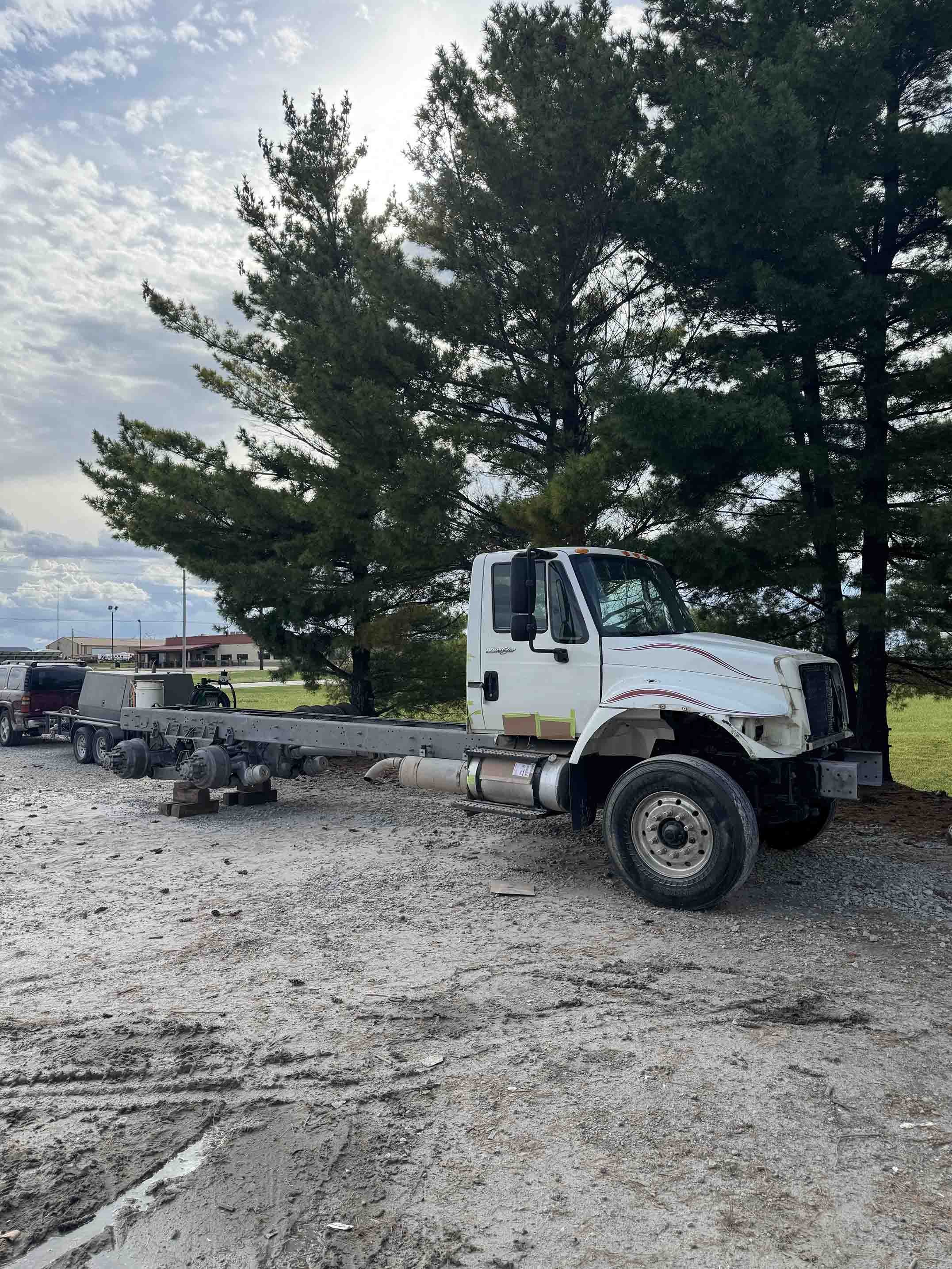 A white flatbed truck without a trailer is parked on a gravel surface next to green trees, with a cloudy sky overhead.