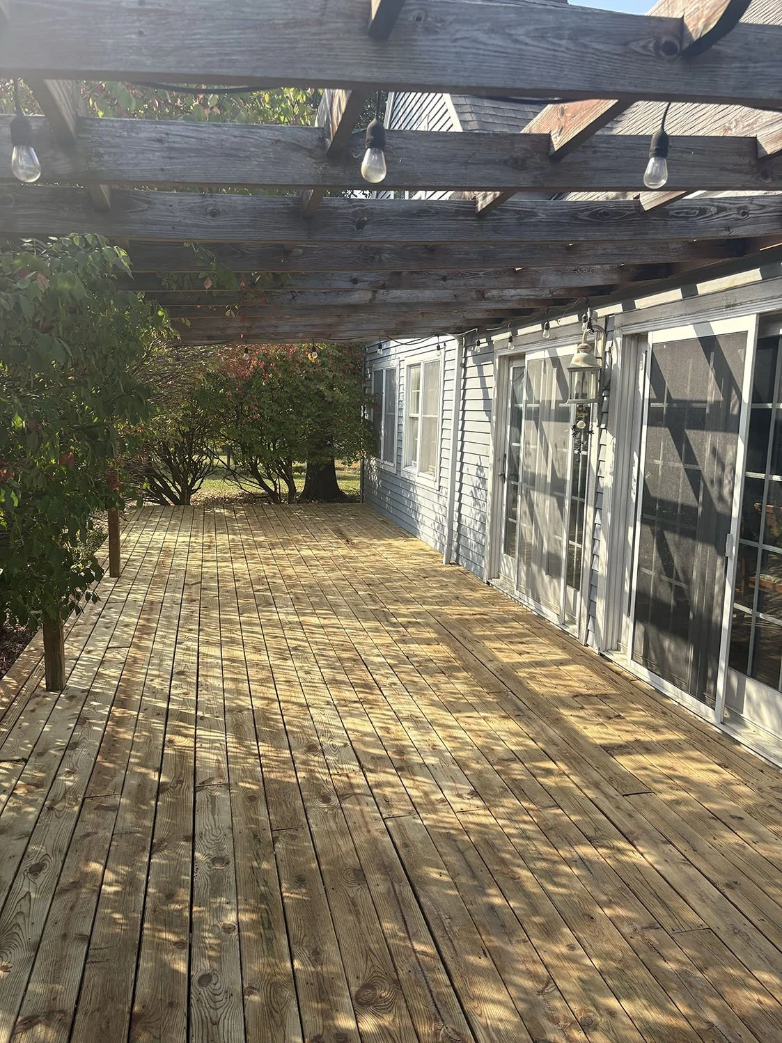 Wooden deck with lattice overhead, string lights, and a house with sliding glass doors and windows. Trees and bushes are visible in the background.