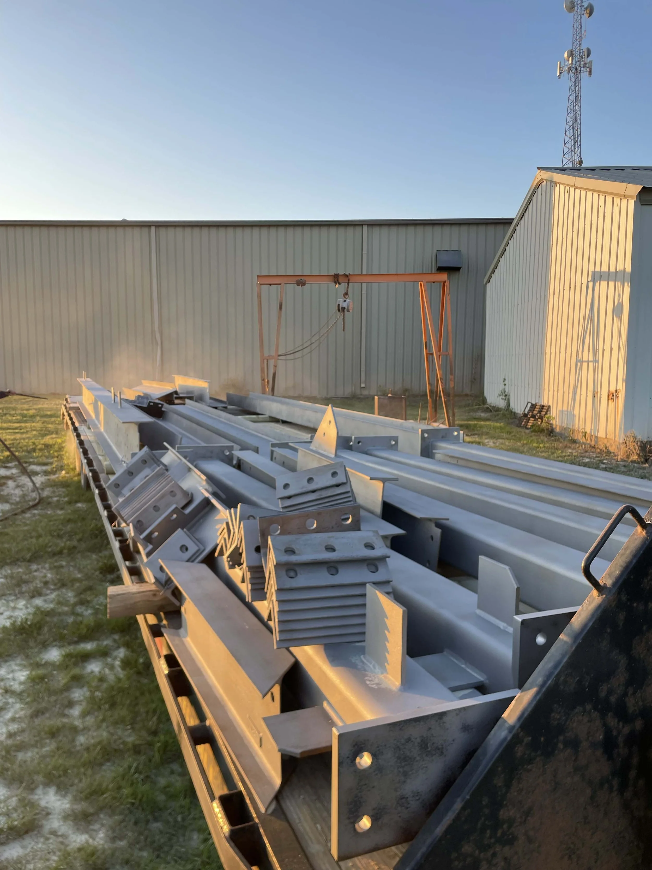 Metal beams and components stored on a flatbed truck outdoors, with a utility building, a crane and a communications tower in the background during sunset.