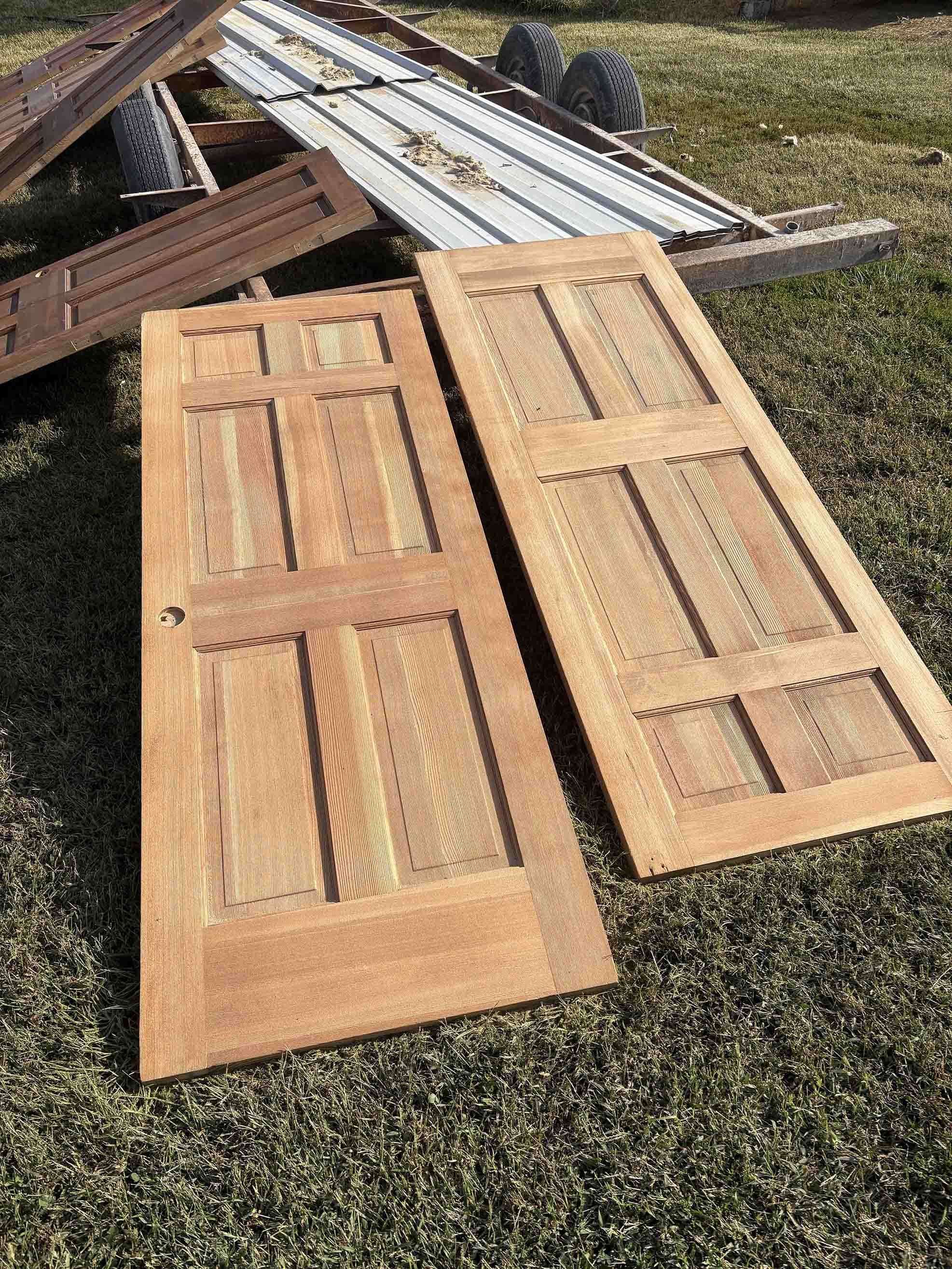Two wooden doors with six panels each, lying on grass, nearby is a trailer with metal sheets and tires.