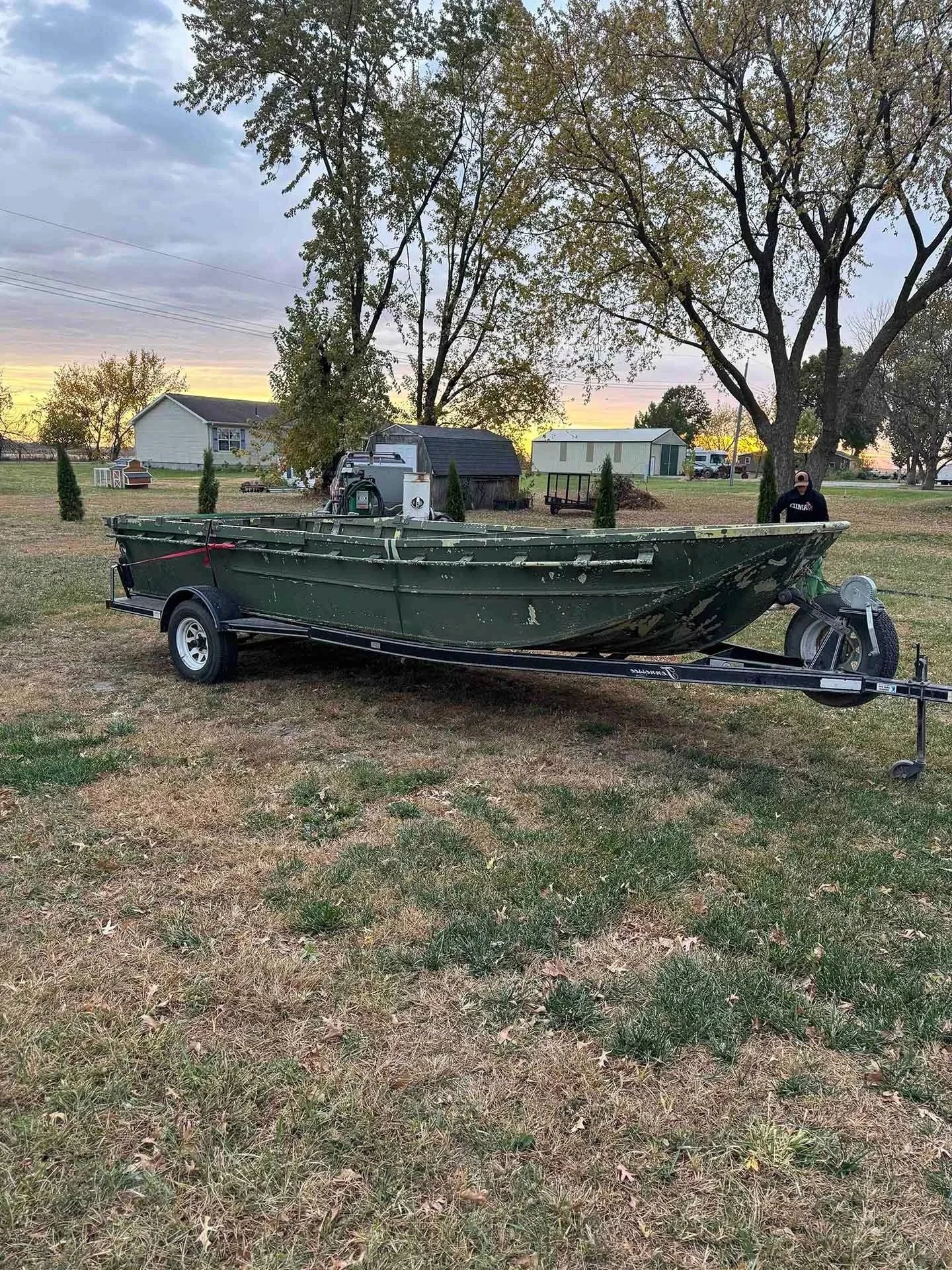 A green boat on a trailer parked on grass with trees, small buildings, and a person in the background at sunset.