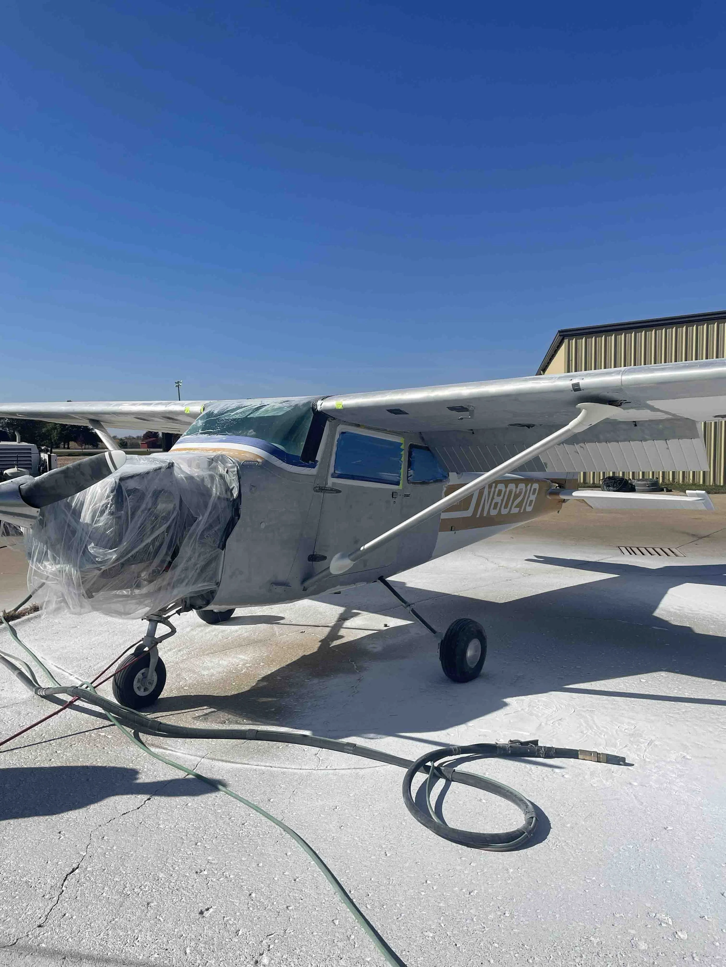 Small airplane on tarmac, covered parts, connected to ground power, with hangar and clear blue sky in background.