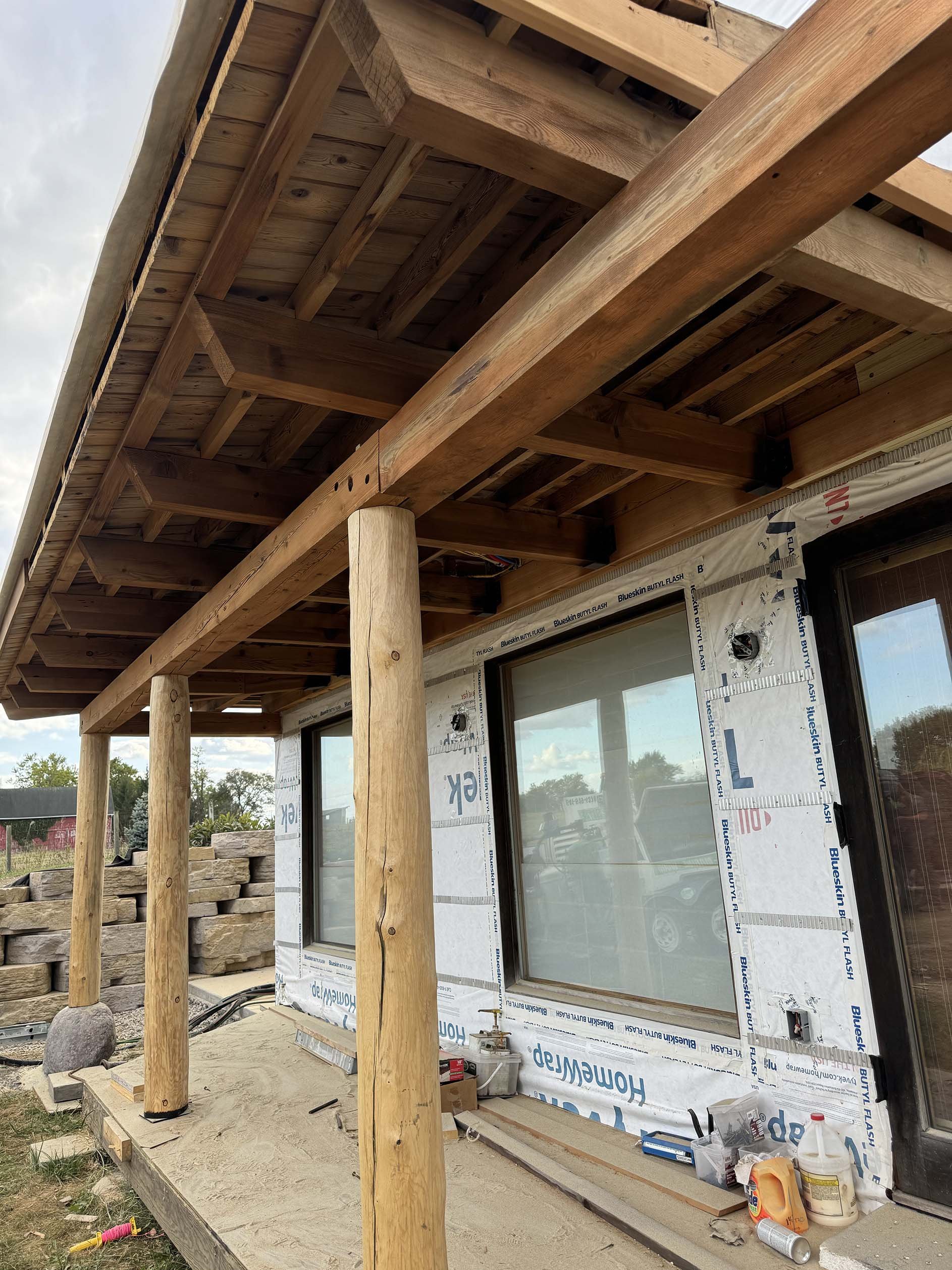 Under construction wooden porch with vertical support beams and a view of a house exterior with new windows, construction tools, and building materials.