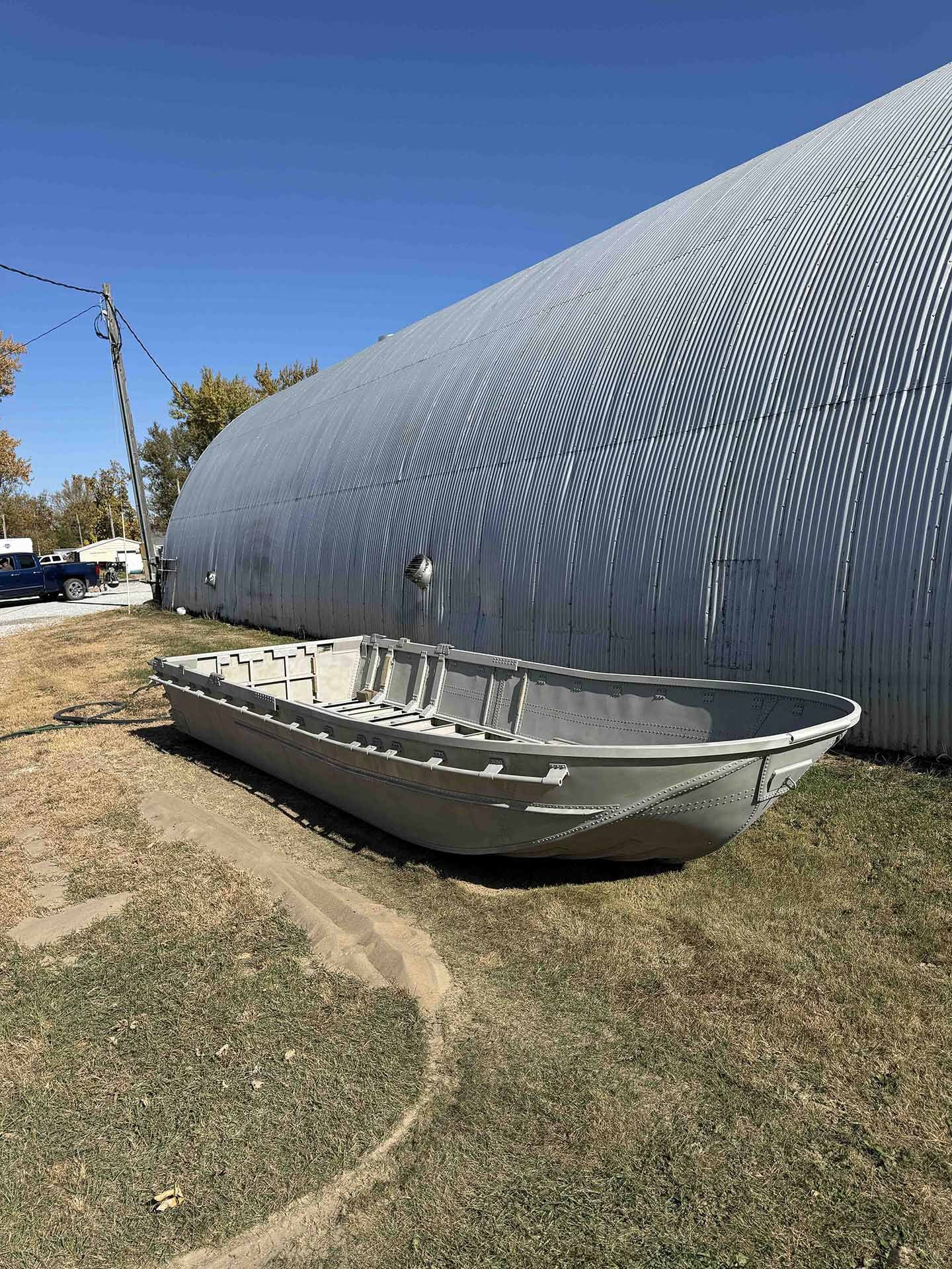 An empty gray boat on the ground outside next to a large metal shed under a clear blue sky.