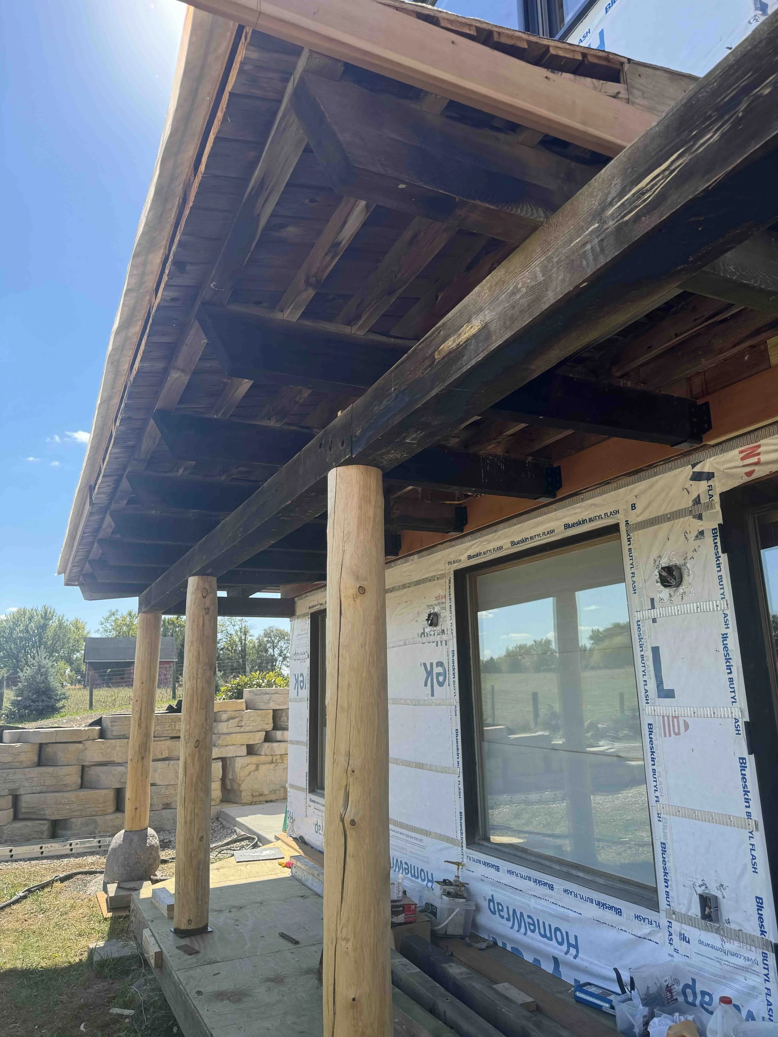 Construction of a house with wooden beams supporting an overhanging roof, siding still in progress, windows installed, and building materials scattered around.