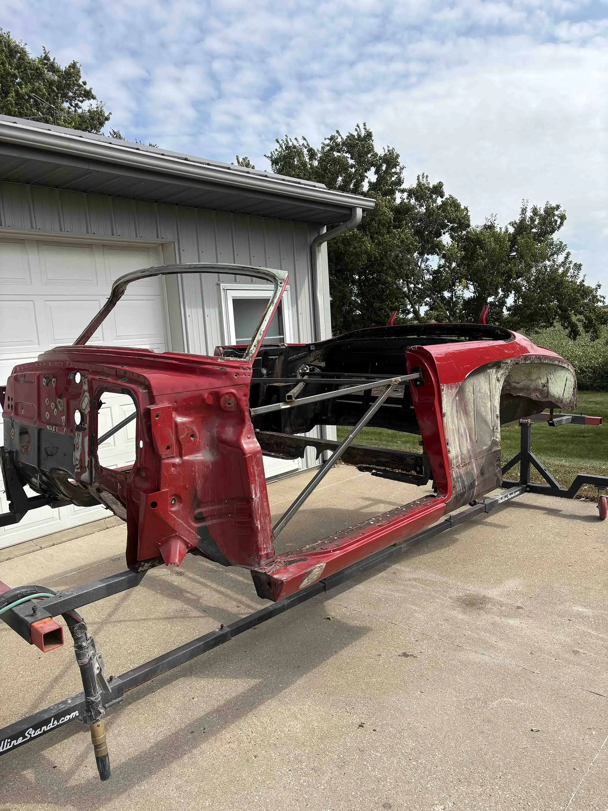 Red car frame on a stand outside near a house with a white garage door, under a partly cloudy sky.