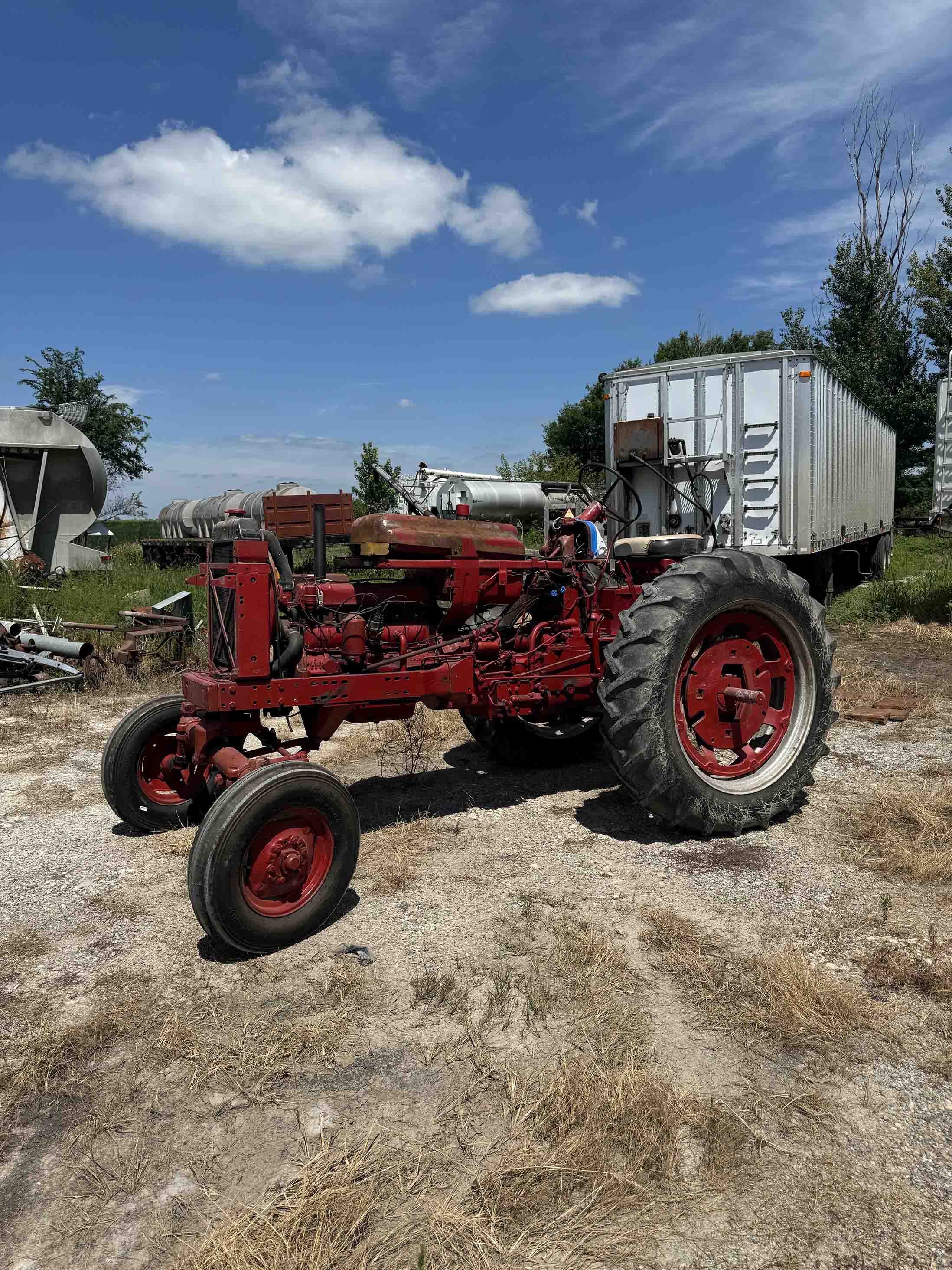 An old red tractor with large rear tires and smaller front tires is parked on a dirt lot with dry grass. In the background, there are metal farm tanks, a silver trailer, and trees under a blue sky with white clouds.