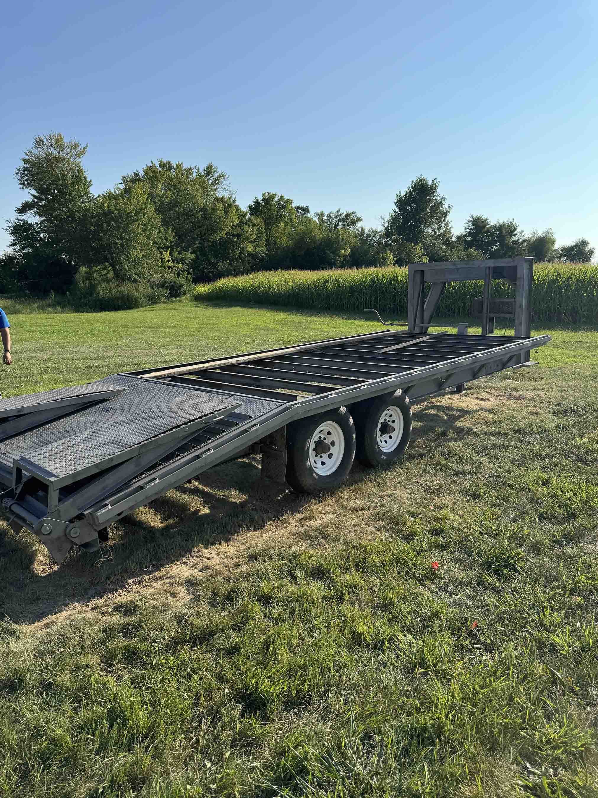 Empty flatbed trailer parked on grassy field with trees and cornfield in the background.