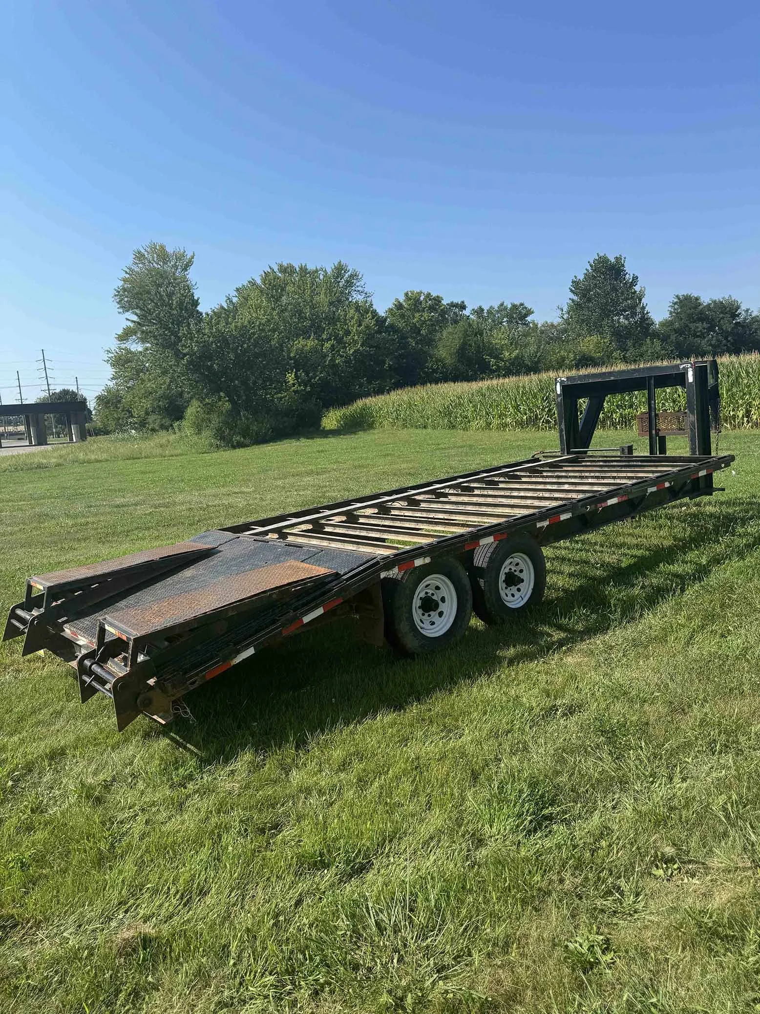 A black flatbed trailer with dual wheels parked on a grassy field under a clear blue sky.