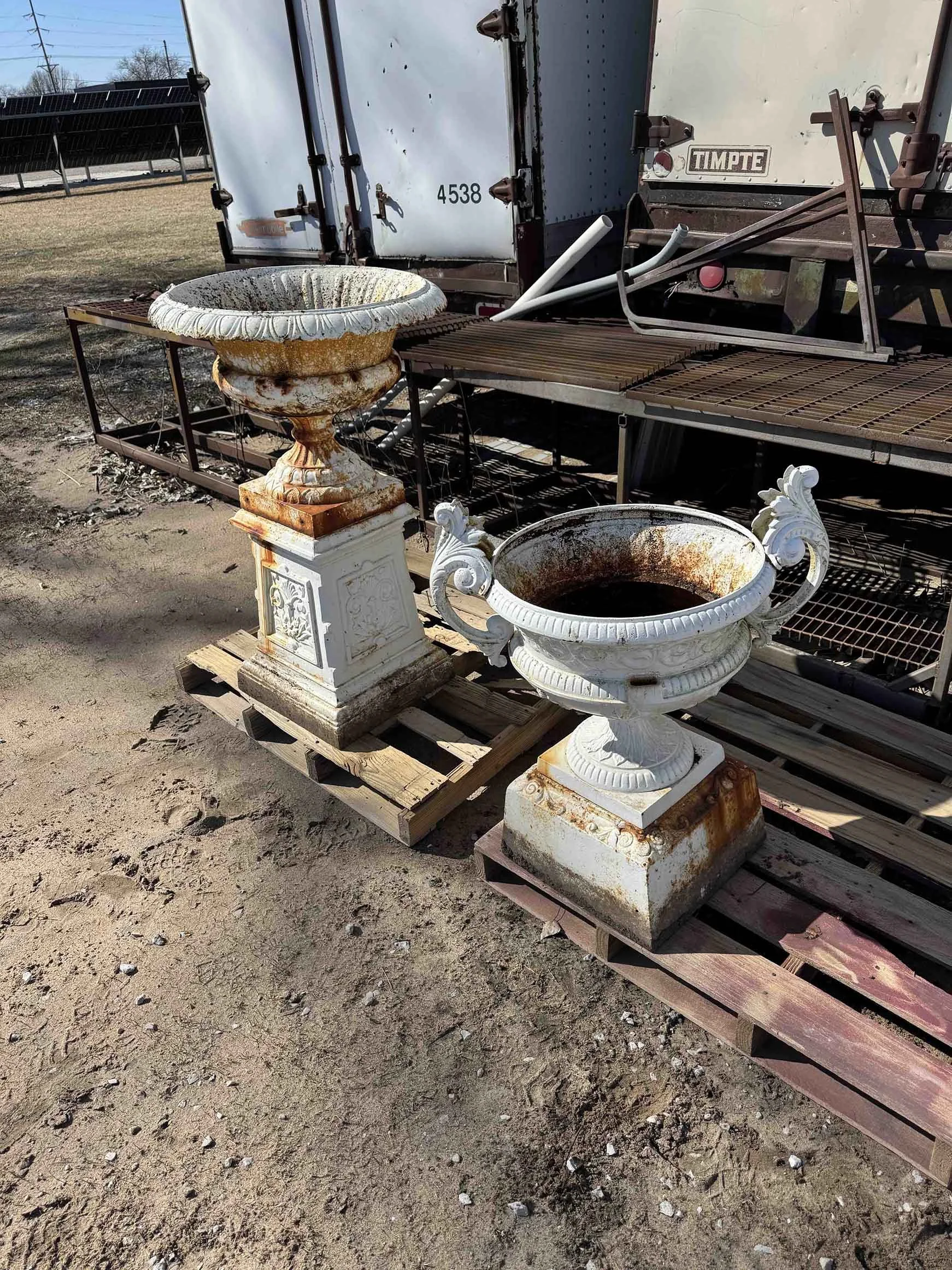 Two ornate, weathered, white decorative urn planters, one larger and one smaller, with rust stains, placed on wooden pallets outdoors.