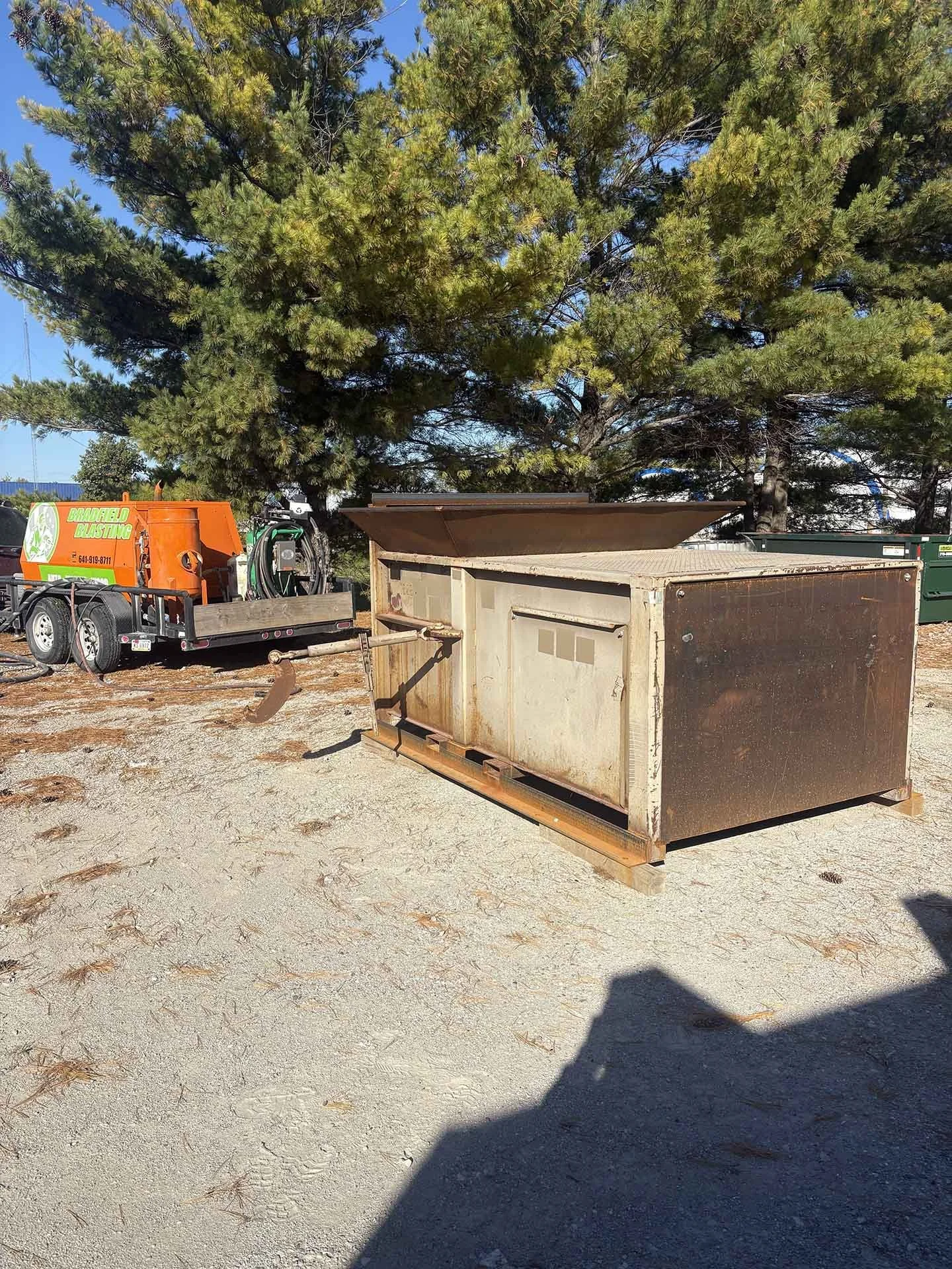 An outdoor scene with a large, rusty, rectangular metal storage container on a wooden pallet. A green tree with dense foliage and a blue sky are in the background, along with a trailer and some equipment on the left side of the image.