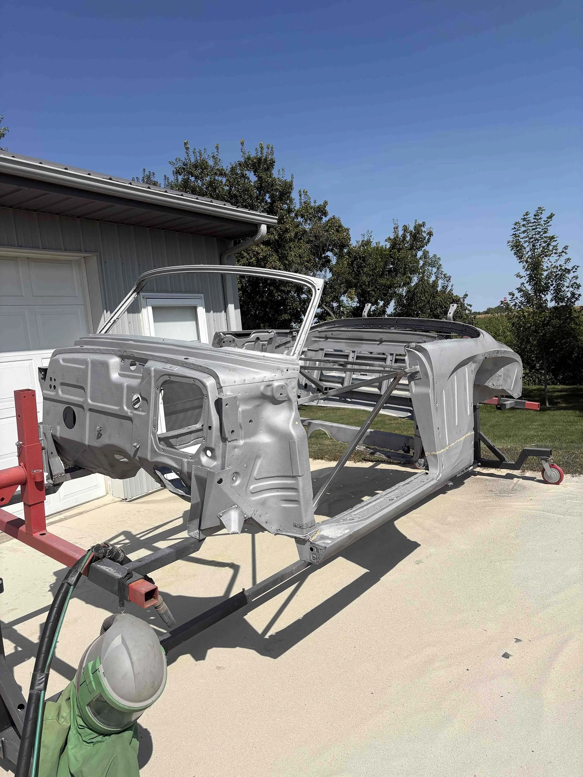 Metal car body shell on a stand outdoors, with a garage and trees in the background.