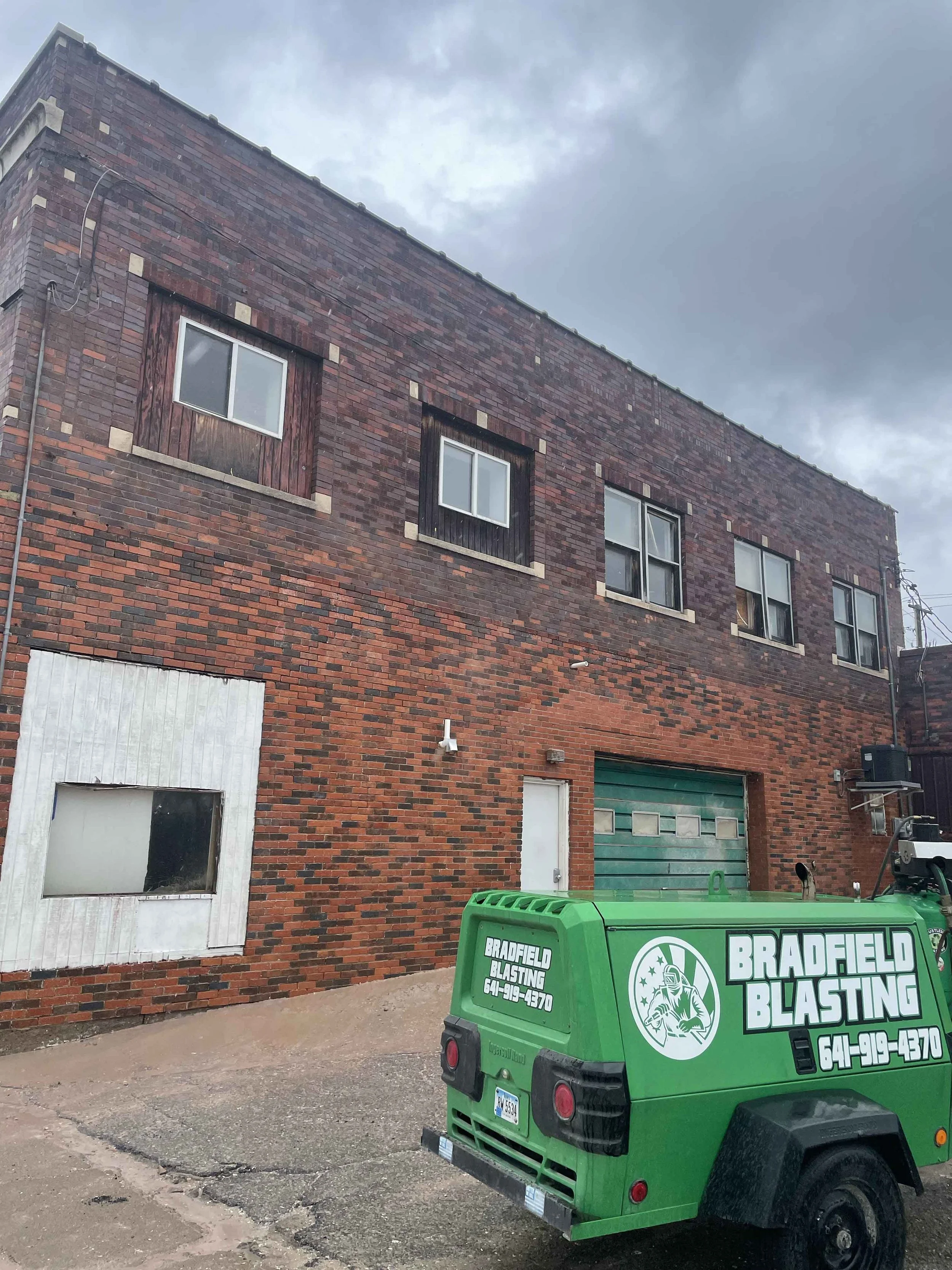 A red brick two-story building with four windows on the second floor, some of which are boarded up. There is a white door and a green garage door on the ground level. A green blasting service vehicle labeled 'Bradfield Blasting' is parked in front of the building.