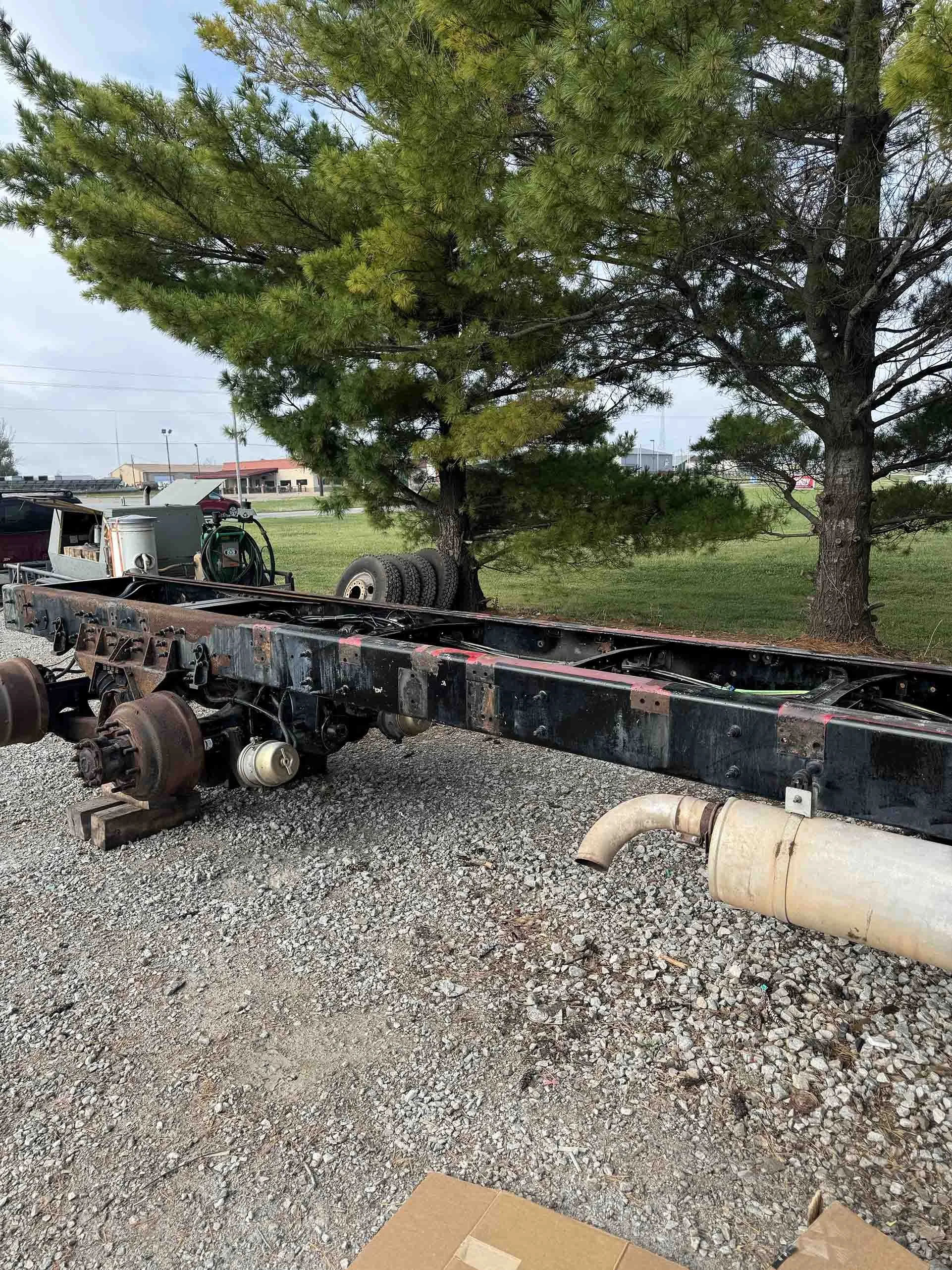 A disassembled truck chassis on gravel ground with two pine trees in the background and a row of tires. The chassis has rusty wheels and a large exhaust pipe extending from it.
