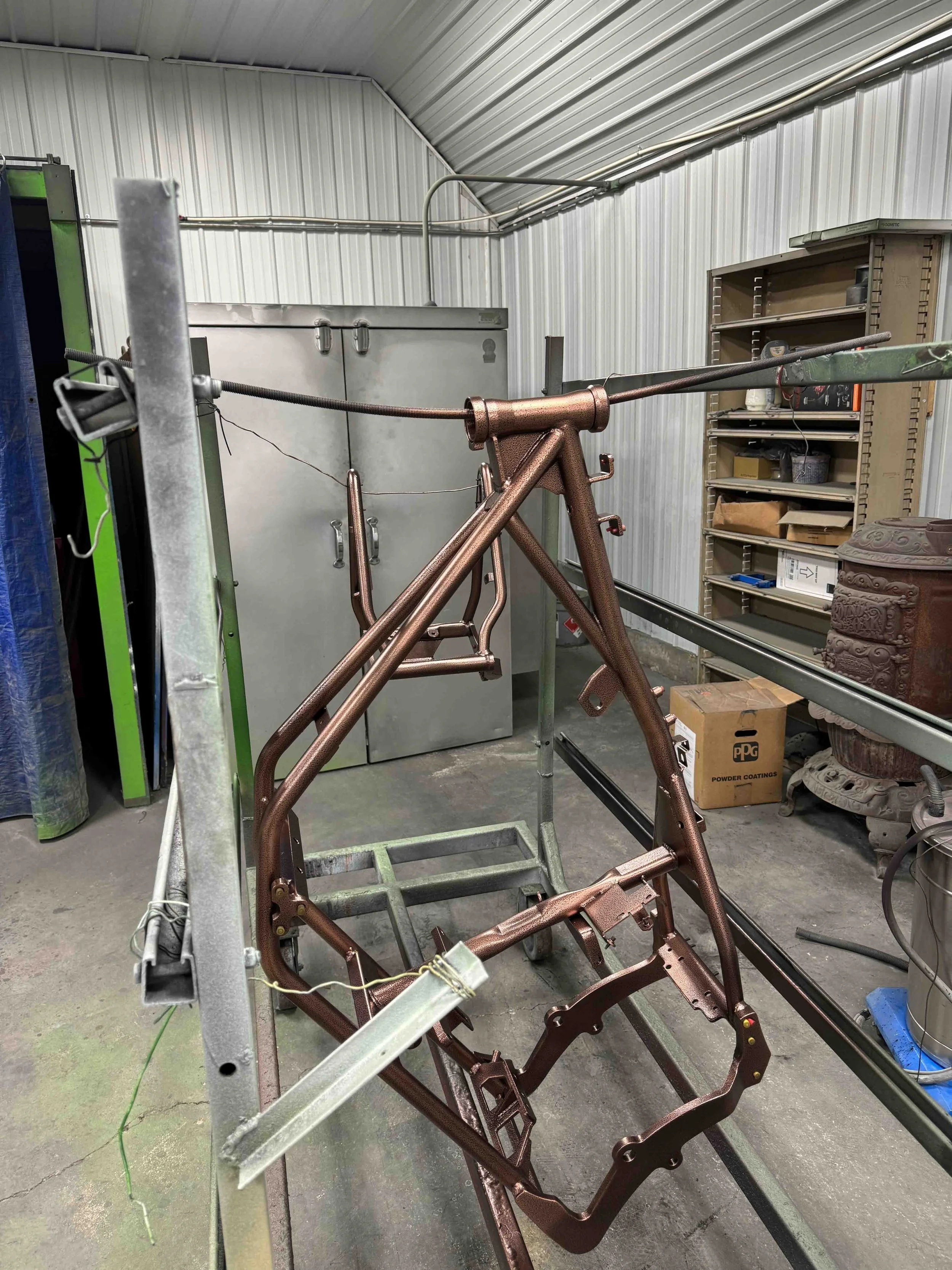 A bicycle frame being painted or coated in a workshop, hanging on a metal stand in front of a storage cabinet and shelving.