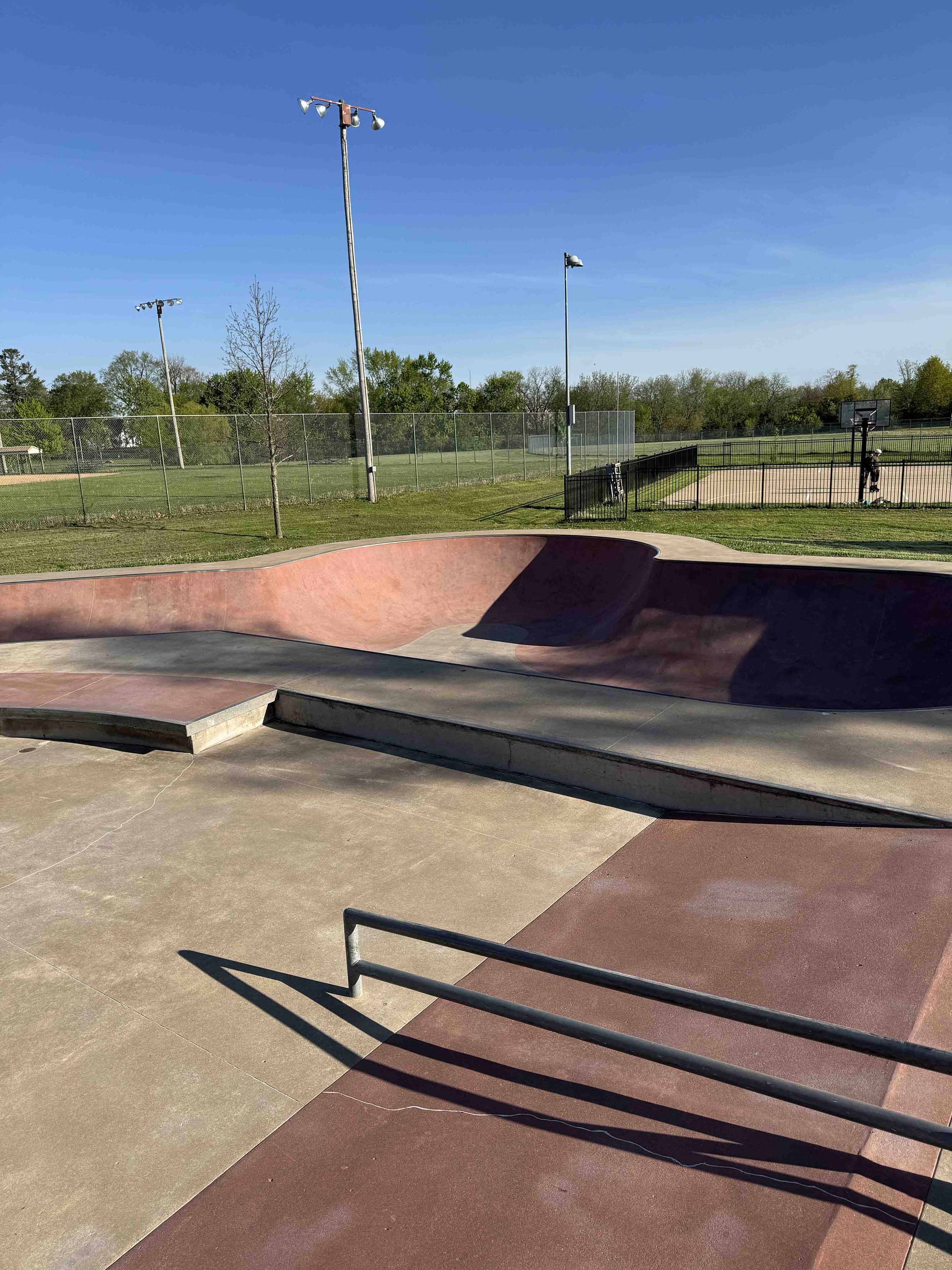 Empty outdoor skate park with ramps, rails, and a basketball court in the background under a clear blue sky.