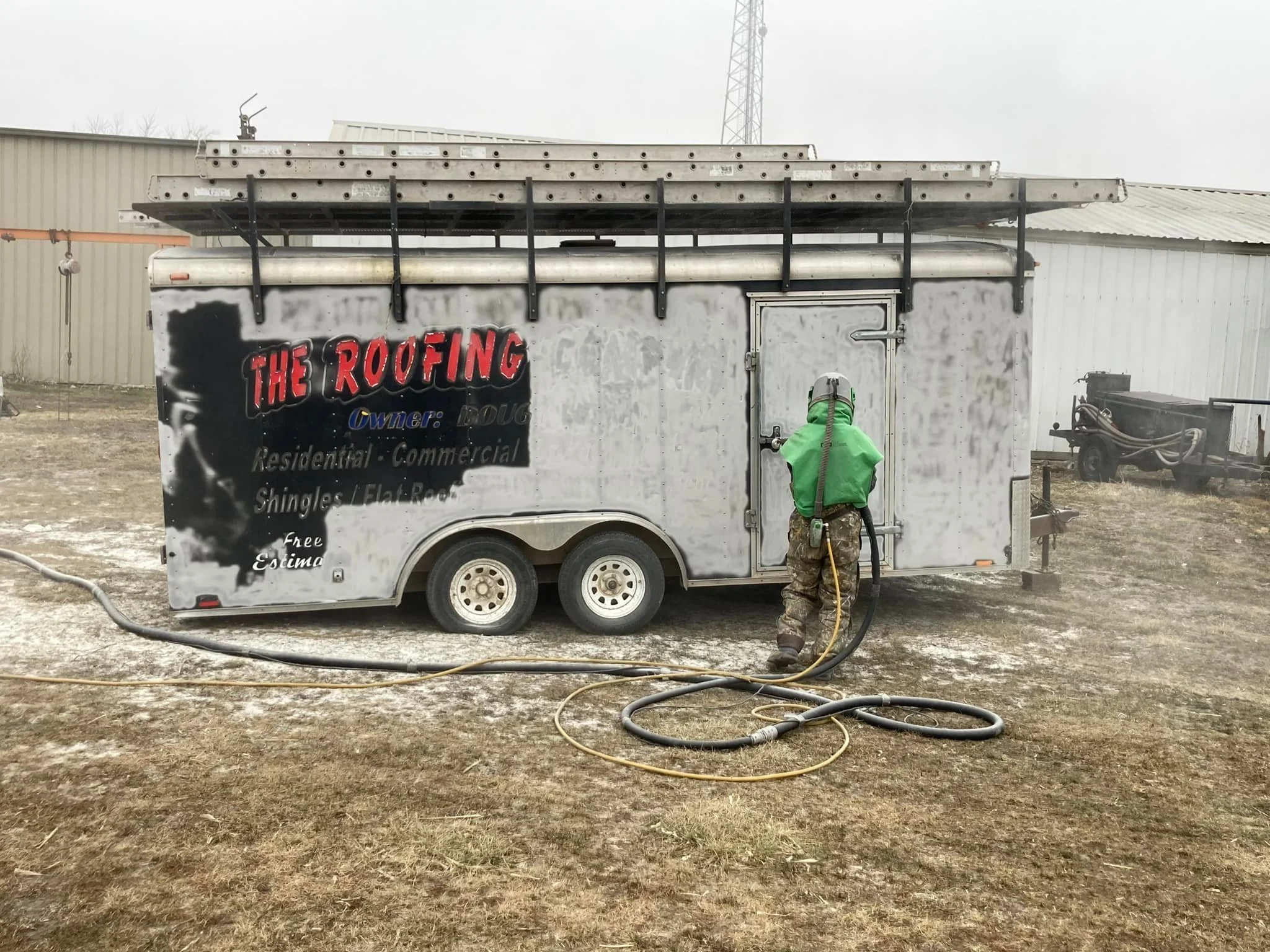 A worker in green protective gear and helmet power washing a mobile roofing business trailer outside on a dirt or gravel lot.