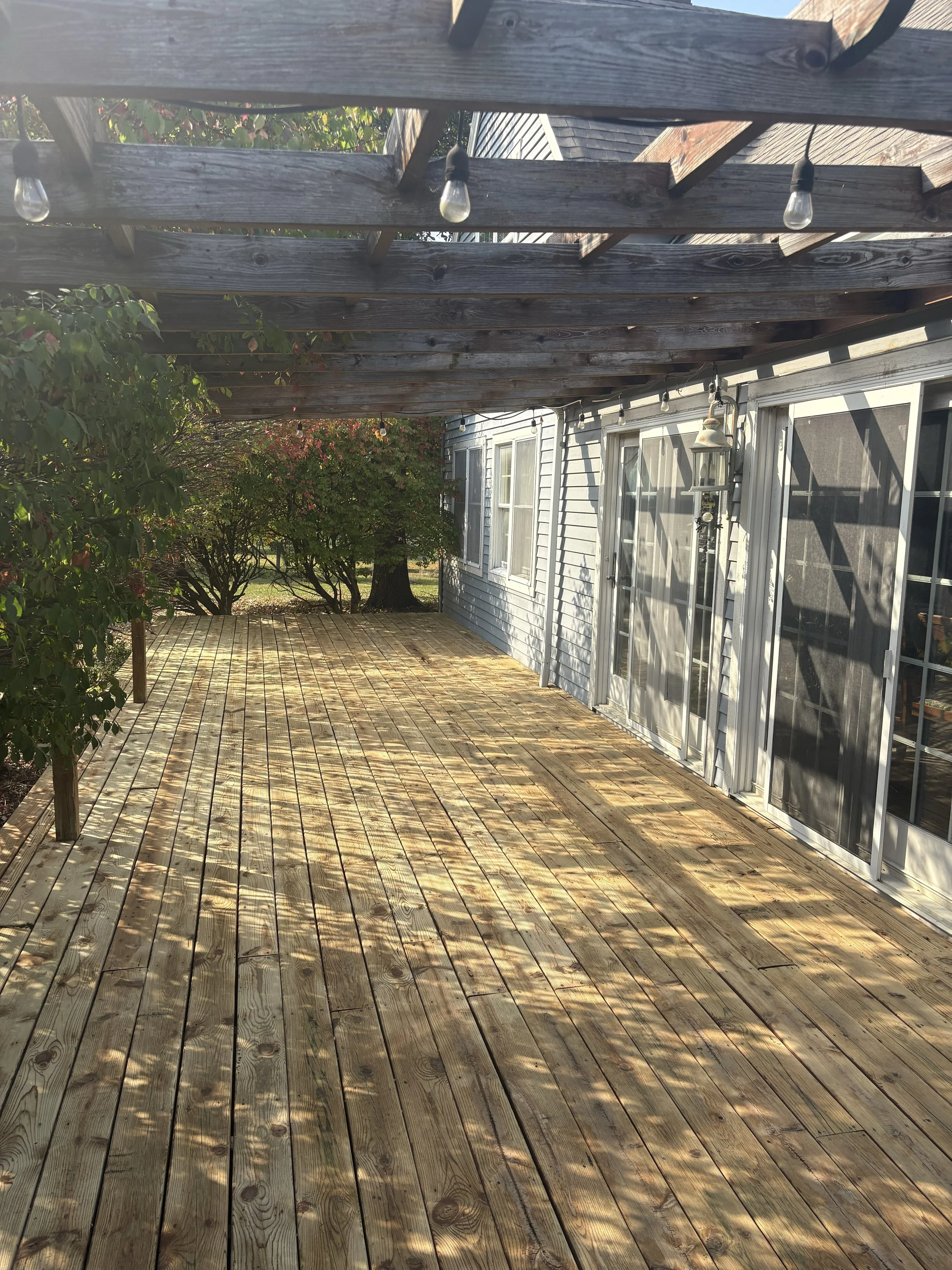 A wooden outdoor deck attached to a house with grass and trees in the background. A pergola with string lights overhead and shadows cast onto the deck.