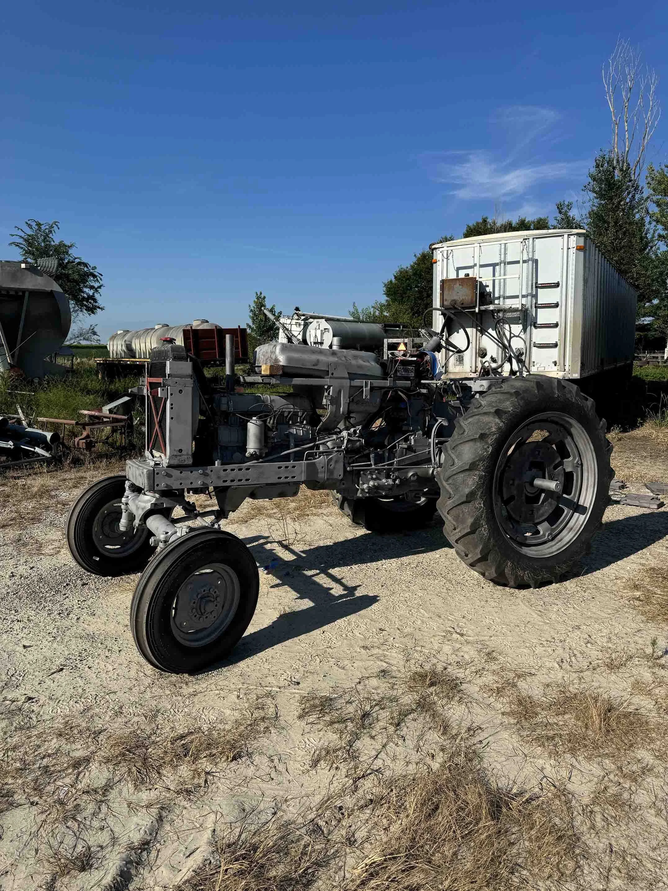 A rusty tractor with a white cargo container attached to its back is parked on a dirt lot with dry grass. The tractor is missing its engine and other parts, and the background includes trees, fuel tanks, and a clear blue sky.