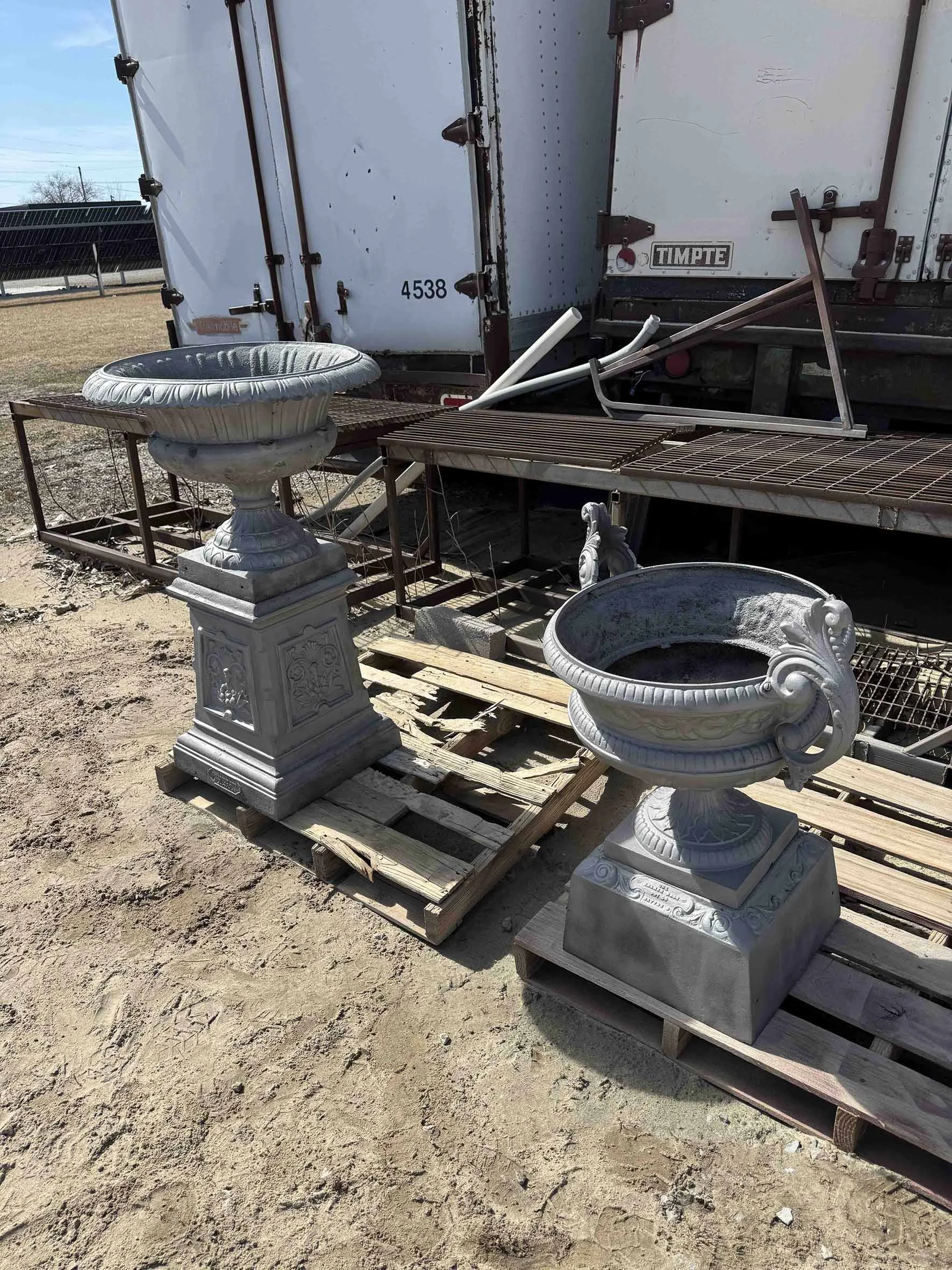 Two decorative stone planters on wooden pallets outside, with metal garden furniture and a trailer in the background.
