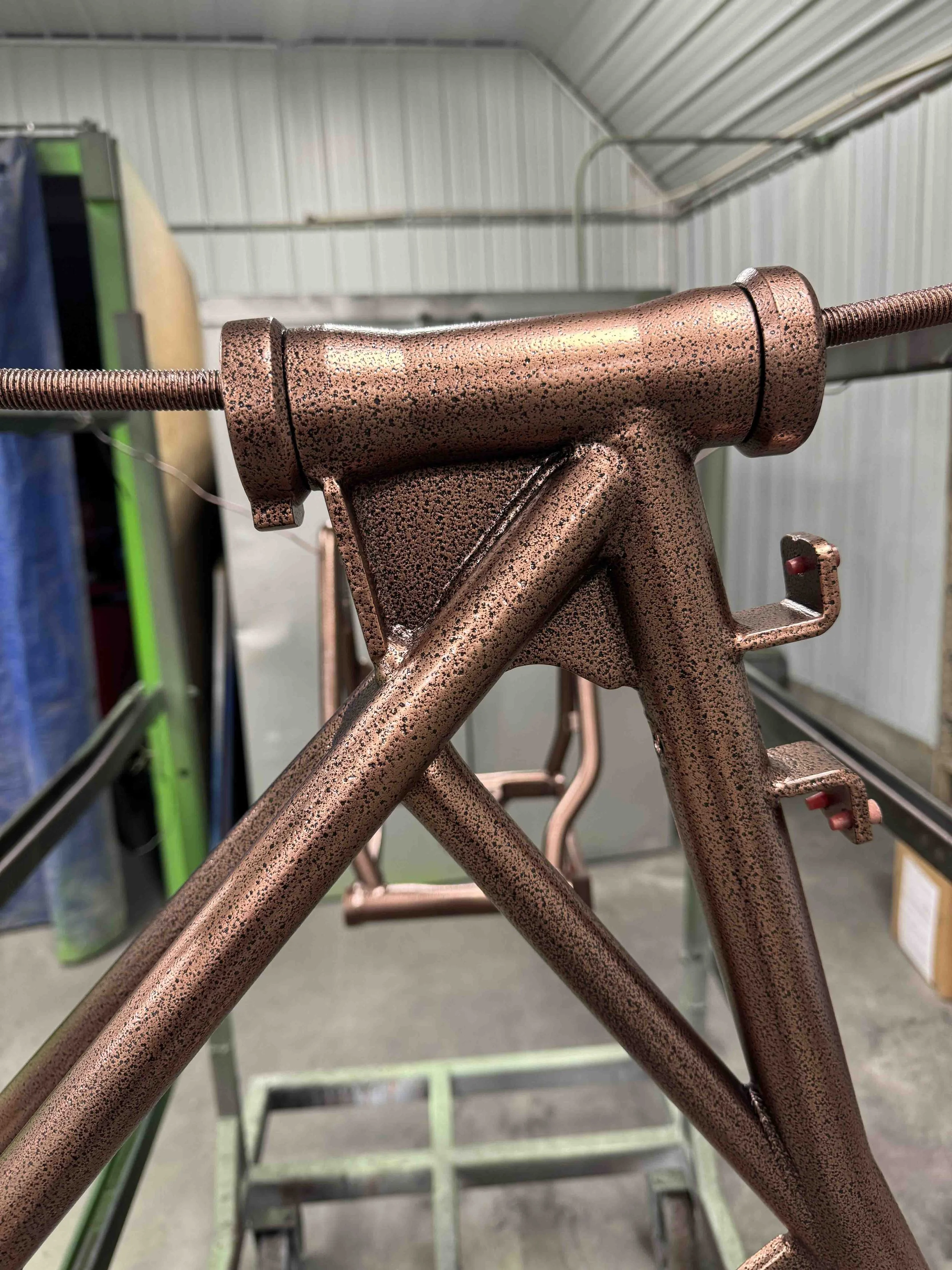 A close-up of a bicycle frame with a speckled, copper-colored finish, mounted on a stand in a workshop.