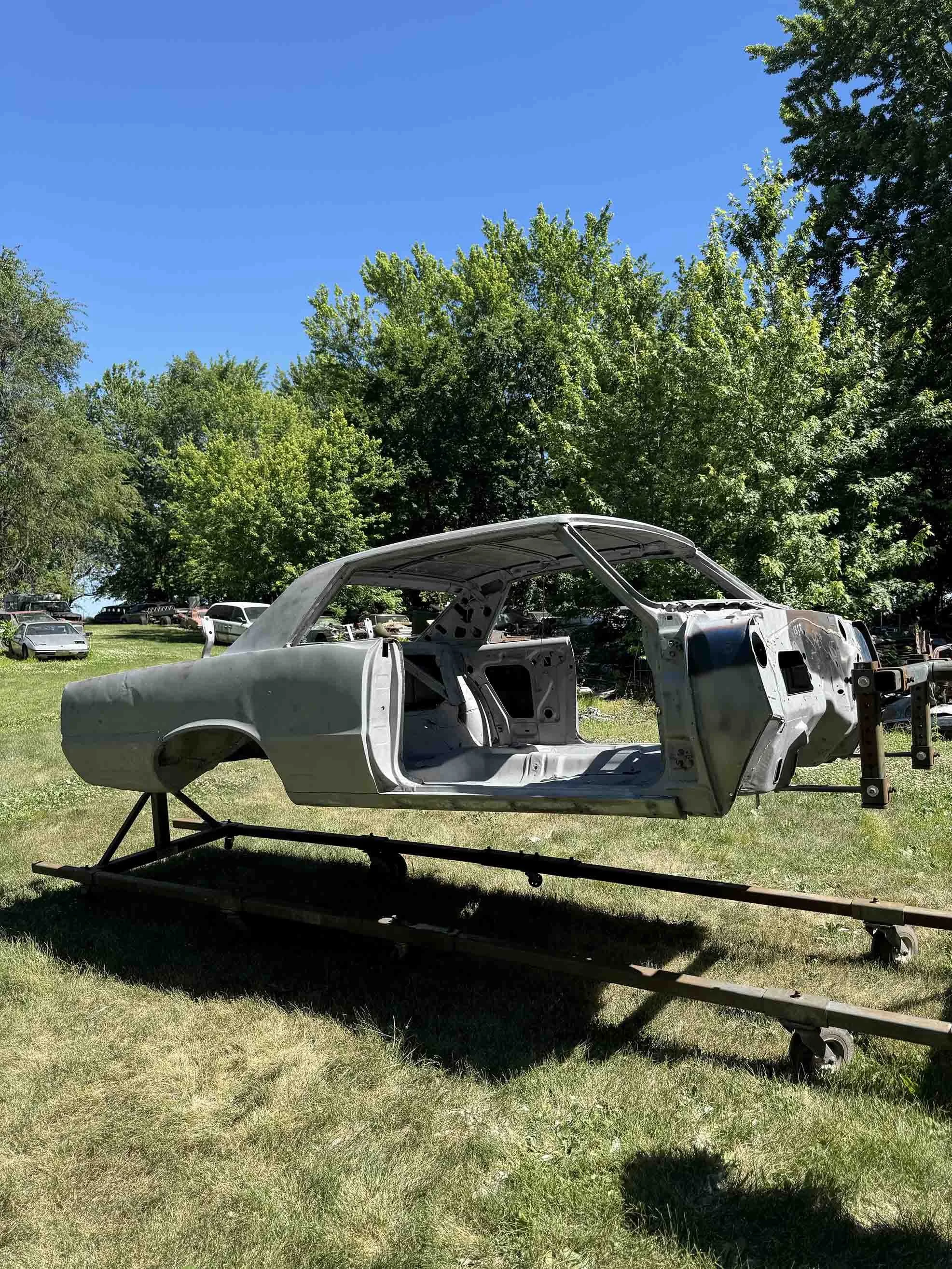 An old car body shell on a rolling platform outdoors, with trees and other vintage cars in the background, under a clear blue sky.