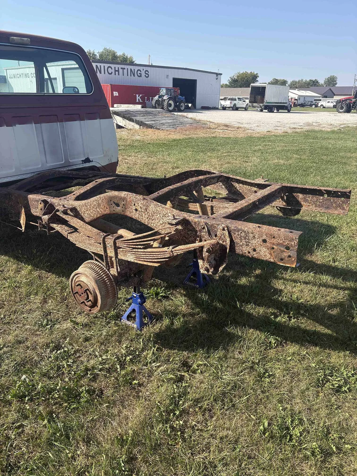 Rusty vehicle chassis with no wheels, supported by blue jackstands on grassy ground, with an old truck and a building in the background.